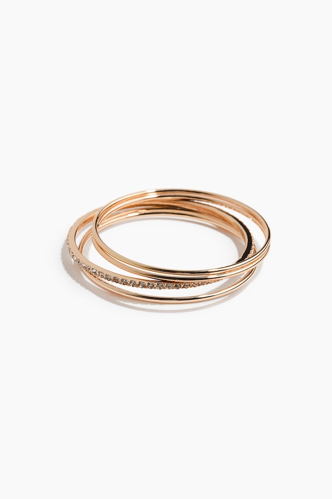 A set of gold-toned bangle bracelets is lying in an overlapping stack, featuring one band embellished with a row of small, clear crystals and three smooth, polished bangles. - 1