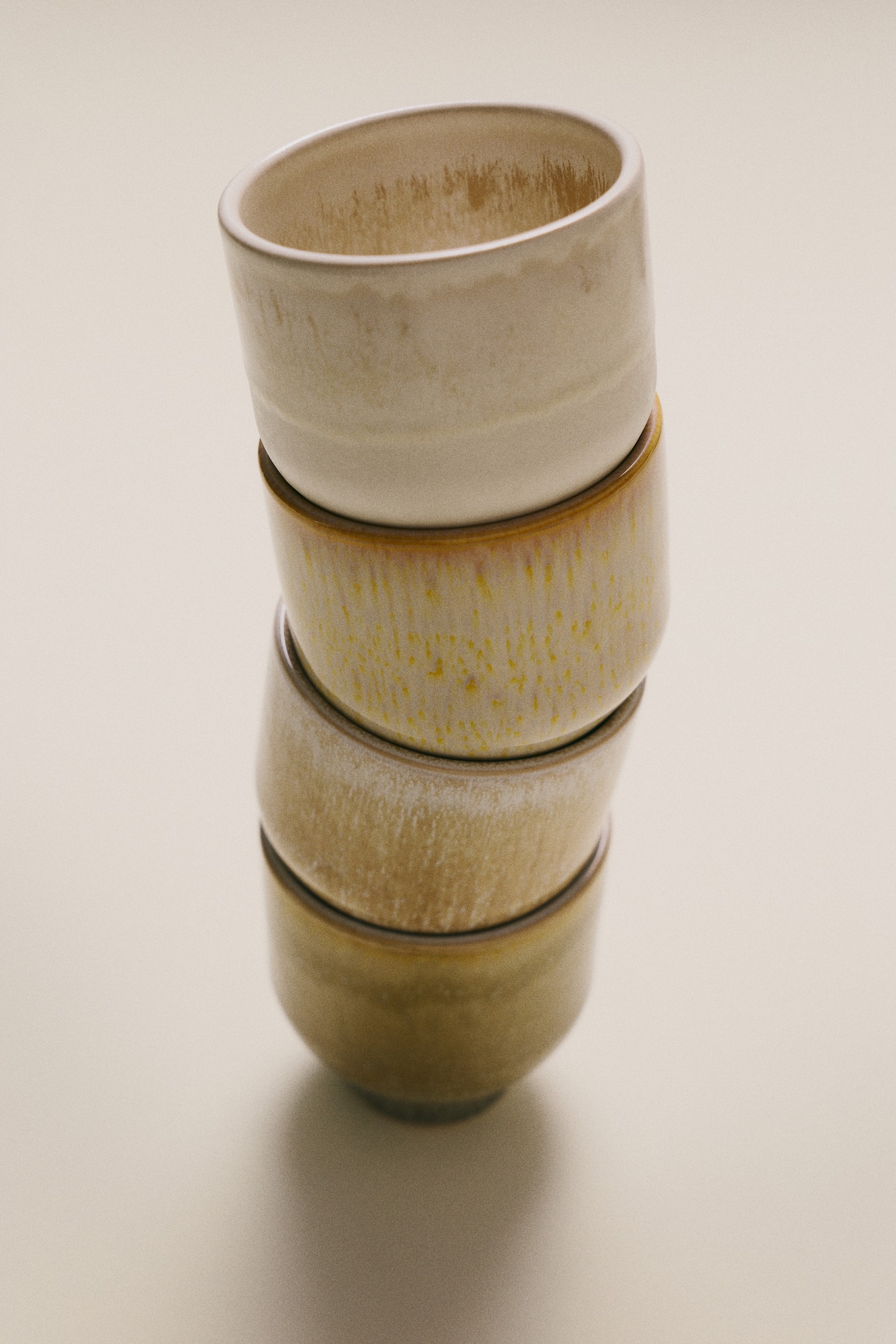 Four handleless ceramic cups are arranged in a horizontal row, facing forward, showcasing a rounded shape that tapers to a narrow foot. Each cup features a distinct earth-toned glaze, ranging from speckled olive green and off-white with a dripped rim to textured beige and pale peach. - 1