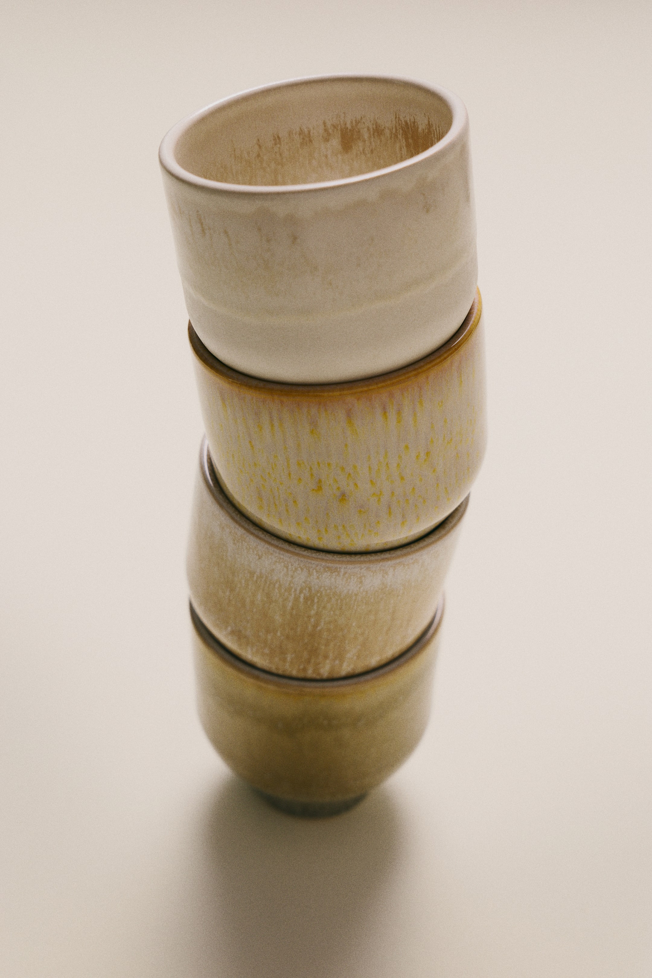 View larger image: Four handleless stoneware mugs with footed bases are resting in a slightly tilted vertical stack, showcasing their unique reactive glaze finishes. The mugs display a range of earthy tones, transitioning from cream at the top to speckled yellow, textured beige, and olive green at the bottom.