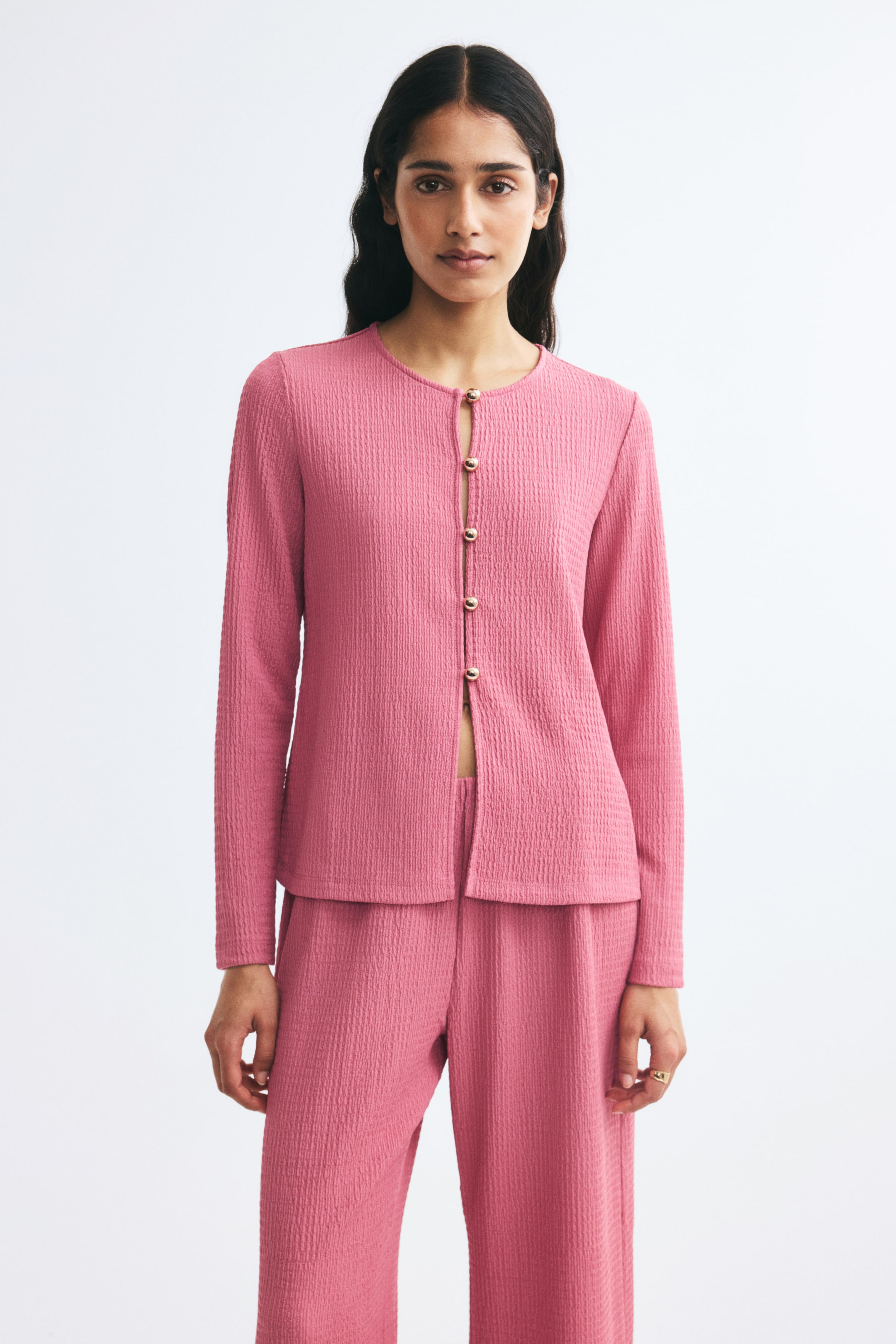 View larger image: A front view shows a pink, fitted cardigan in textured jersey fabric, featuring a round neckline, long sleeves, and small gold-tone ball buttons down the front. The cardigan is paired with matching trousers in the same textured pink fabric.