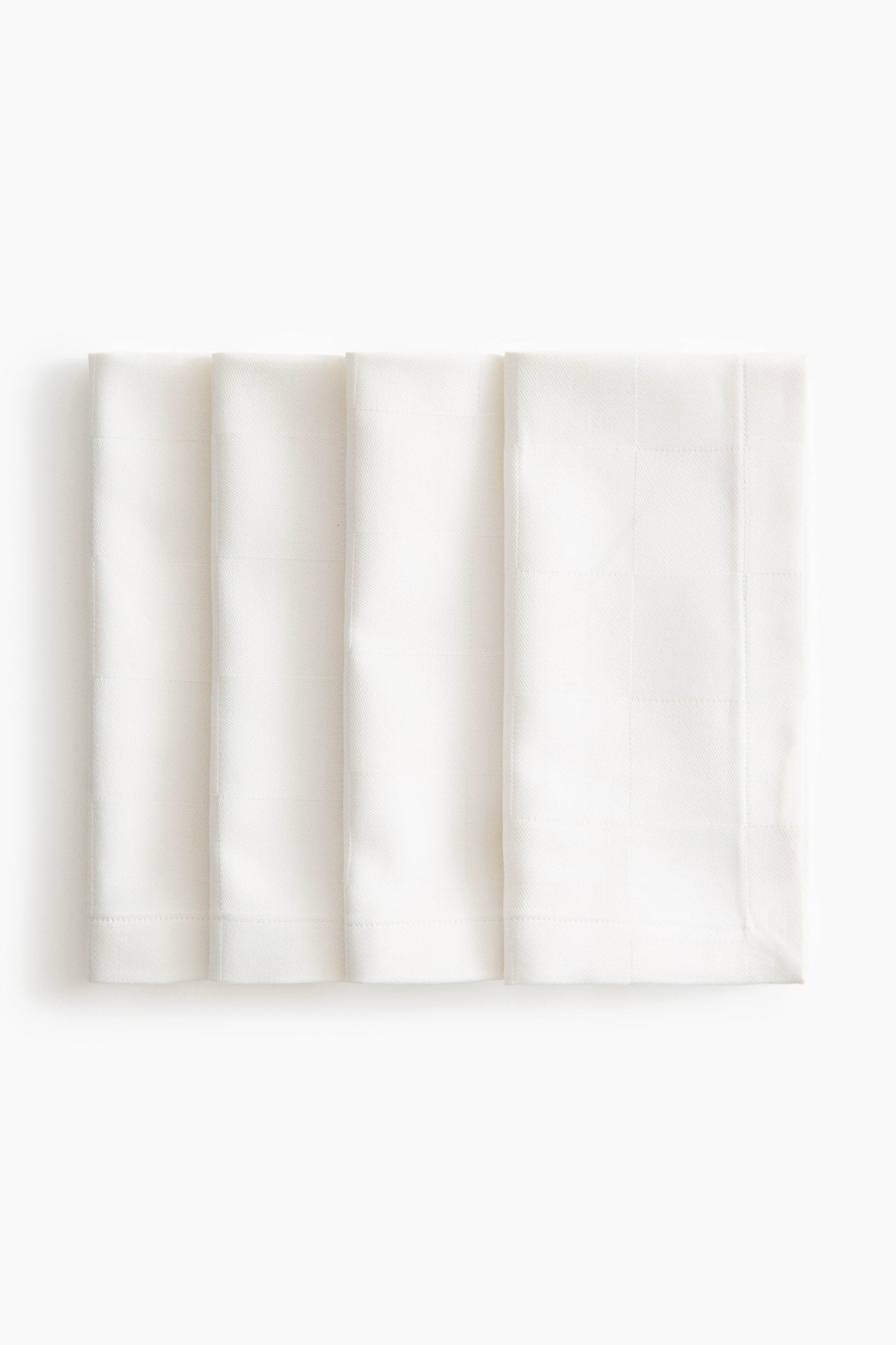 View larger image: Four white cotton napkins are overlapping in a row, facing front, displaying a subtle tone-on-tone woven check pattern and a stitched, folded hem along the bottom edge.