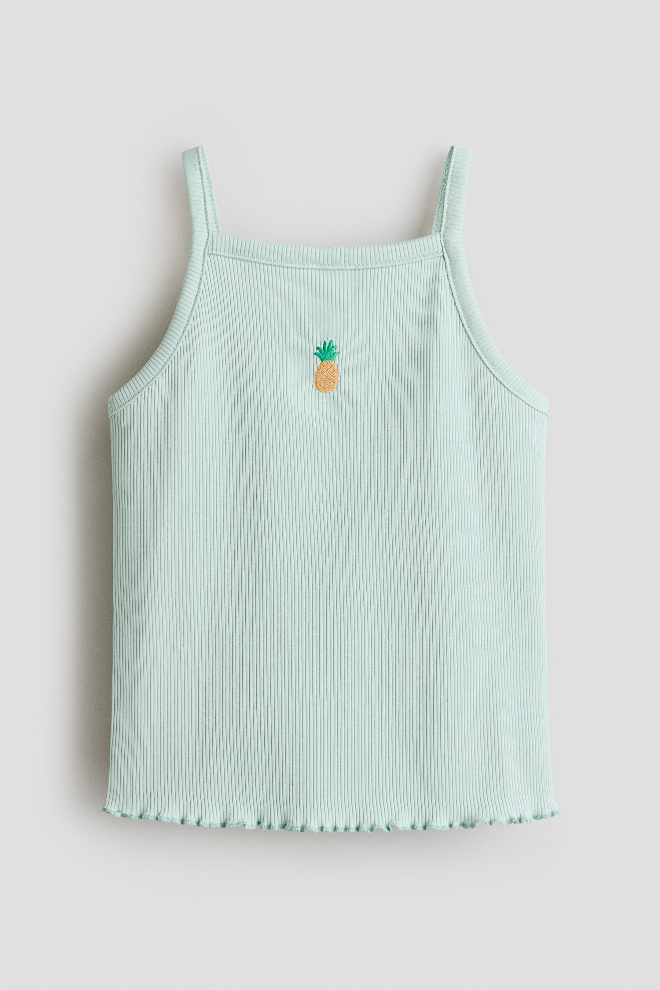 Größeres Bild anzeigen: Ein hellgrünes geripptes Camisole wird in der Vorderansicht gezeigt, mit dünnen Trägern und einem quadratischen Ausschnitt. Ein kleines gesticktes Ananas-Motiv ziert die obere Vorderseite, und der untere Saum endet mit einem gerüschten Rand.