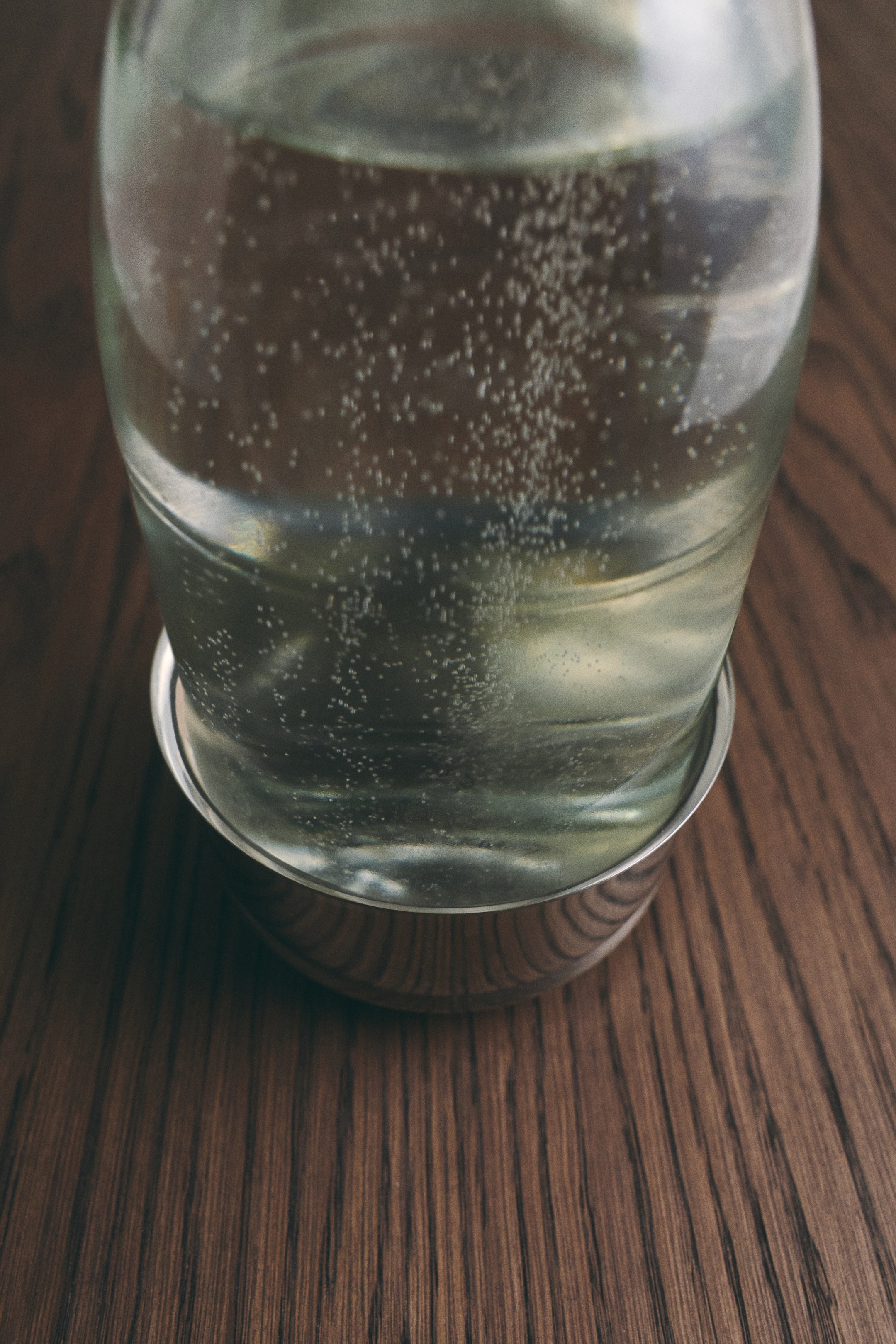 Größeres Bild anzeigen: Von oben betrachtet hält ein polierter Flaschenuntersetzer aus Edelstahl den Boden einer klaren Glaskaraffe mit Sprudelwasser. Der runde Untersetzer hat eine hochreflektierende, metallische Oberfläche mit einem glatten, geschwungenen Rand.