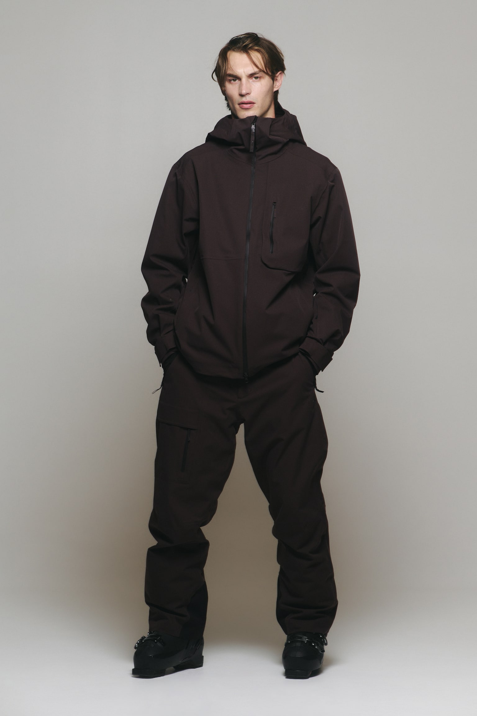 A dark brown technical outerwear suit, consisting of a hooded zip-up jacket and matching loose-fitting trousers, is being worn by the model. The model is standing with hands placed inside the trouser pockets, with the trousers worn over black ski boots. - 1