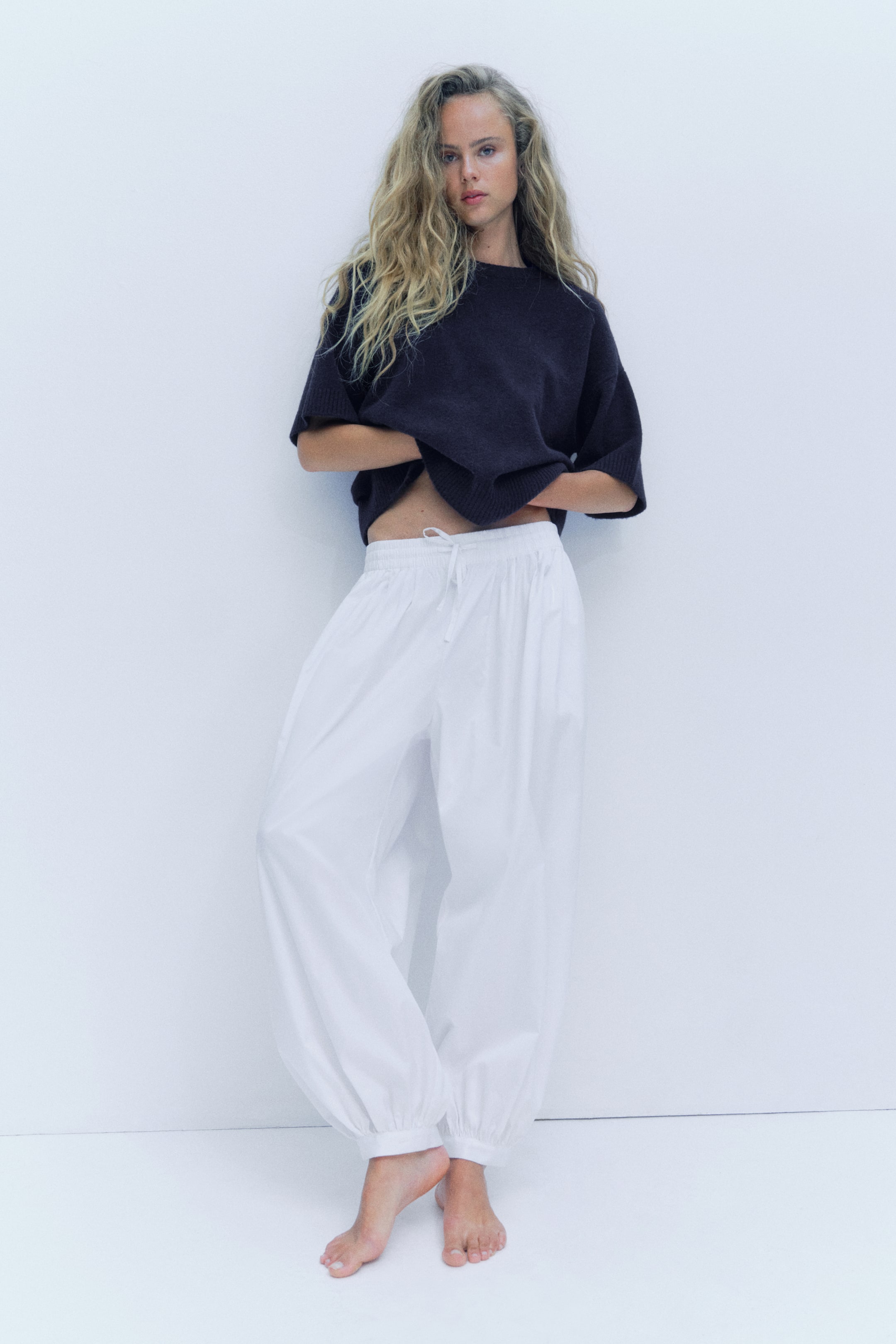 View larger image: An oversized T-shirt in a soft, dark knit is worn by the model, featuring a round ribbed neckline, dropped shoulders, elbow-length sleeves, and ribbing at the cuffs and hem. The model is holding up the T-shirt's hem.