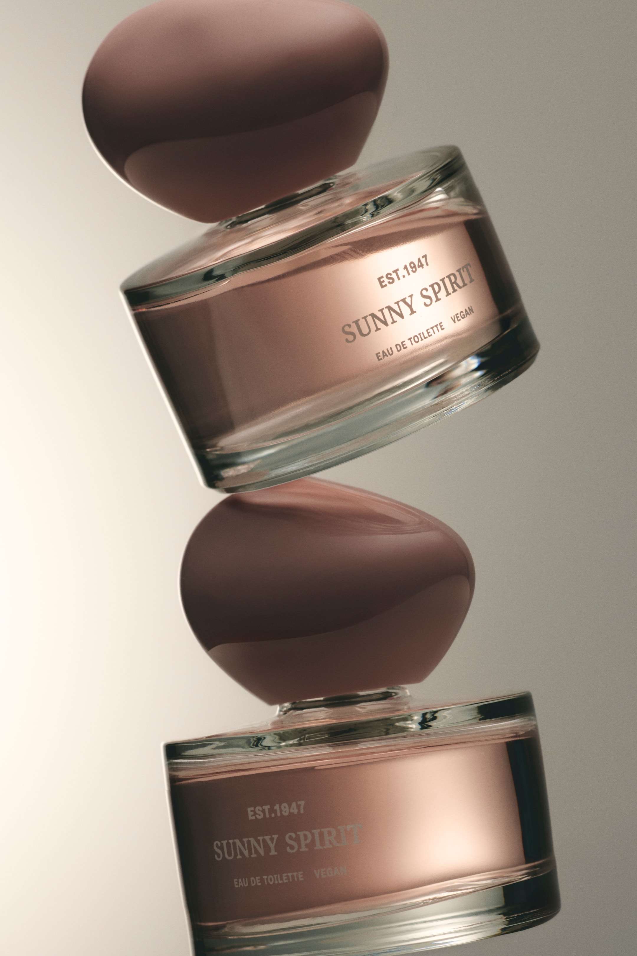 Nagyobb kép megtekintése: Két parfümflakon függőlegesen egyensúlyoz, az egyik fejjel lefelé a másik tetején, mindkettő enyhén jobbra dőlve. Mindkét áttetsző üvegflakon rózsaarany színű folyadékot tartalmaz, tompa szilvalila, szabálytalan formájú kupakkal rendelkezik, és elülső címkéjén szerepel: "EST.1947", "SUNNY SPIRIT", "EAU DE TOILETTE" és "VEGAN".