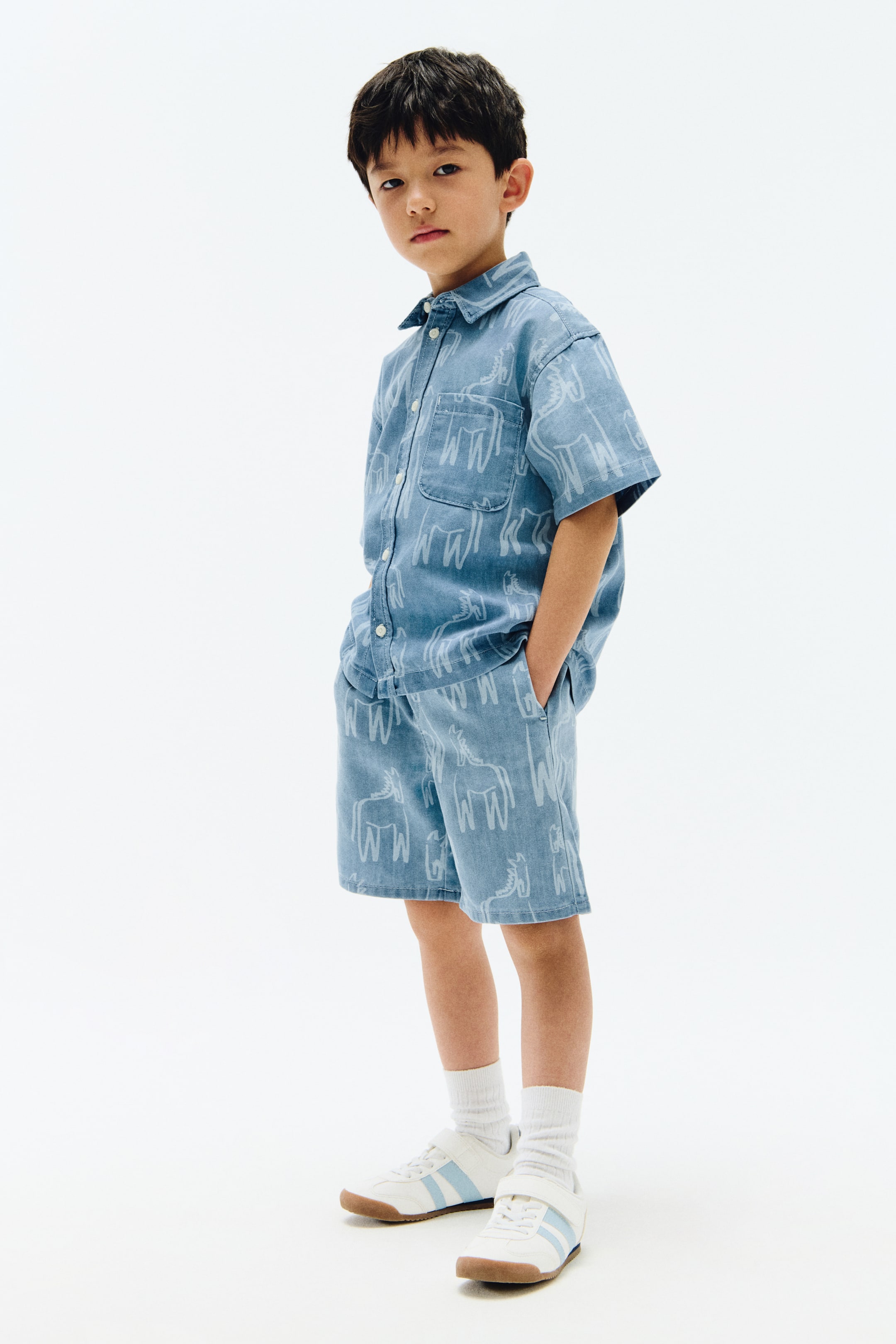 View larger image: A light blue denim short-sleeve shirt and matching shorts set is shown, angled slightly to the left. The set features a repeating pattern of stylised light-coloured horse motifs, with the shirt designed with a collar, front button closure and a chest pocket, while the shorts come with side pockets.