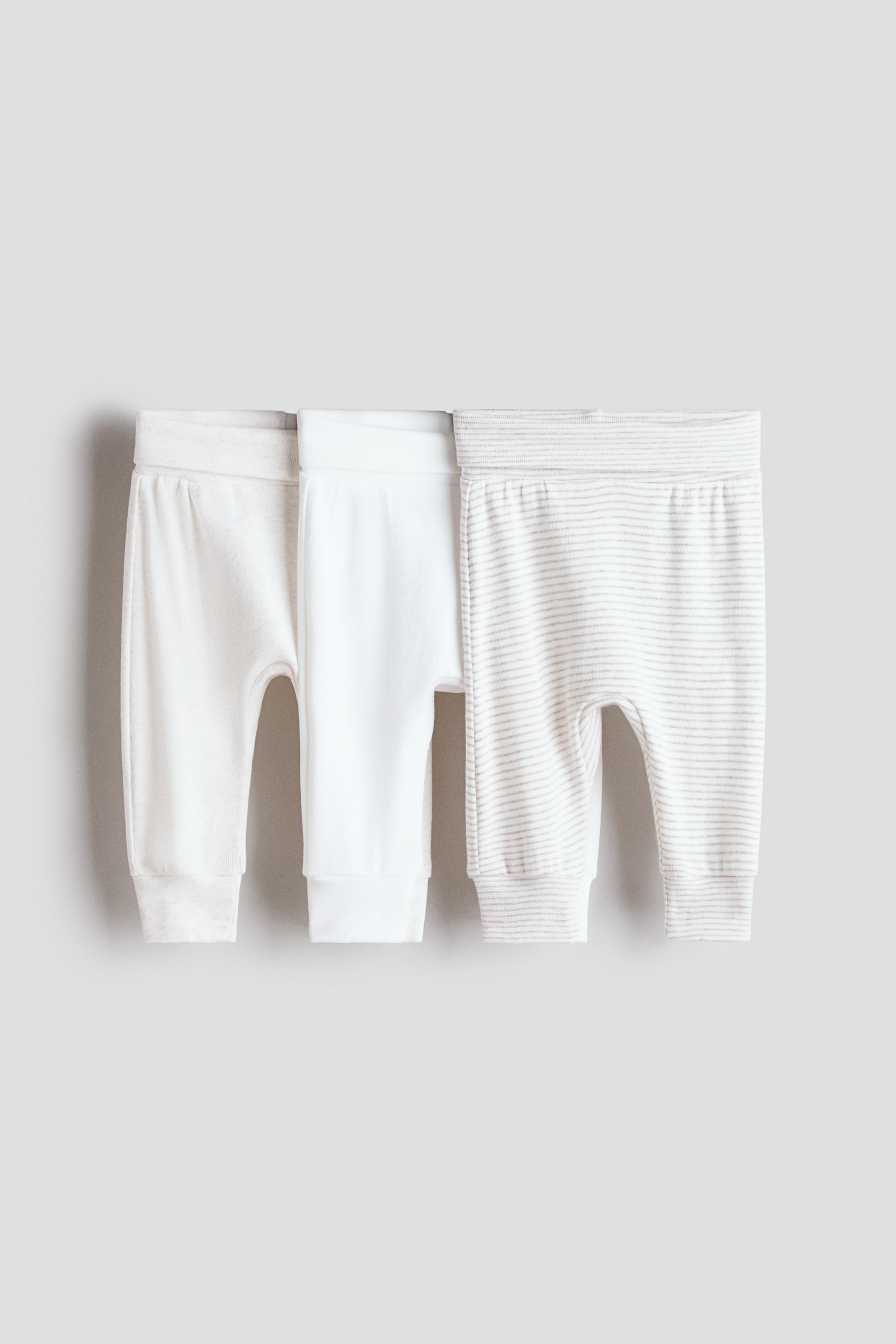 查看大圖: 三件超柔軟棉質針織長褲展示其正面設計，每件均配有寬大的折疊腰帶和束口褲腳。這套裝包括一條淺米色純色長褲、一條純白色長褲，以及一條帶有細橫條紋的淺米色長褲。