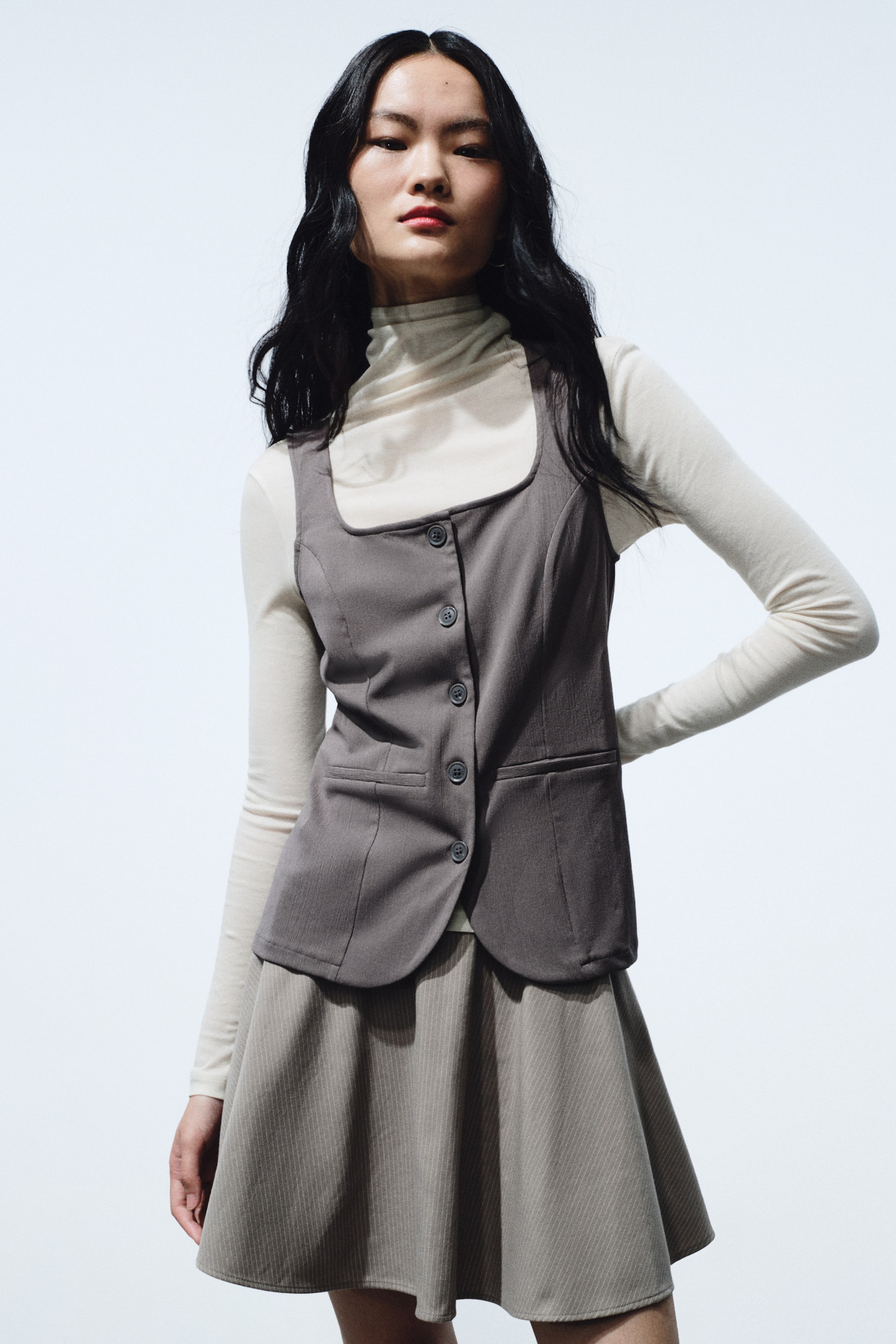 拡大画像を表示: グレーのベストはスクエアネックと5つボタンのフロント開閉が特徴です。フロントにフラップポケットが2つ付き、裾はラウンドヘムで、クリームのタートルネックの上に重ねて着用しています。