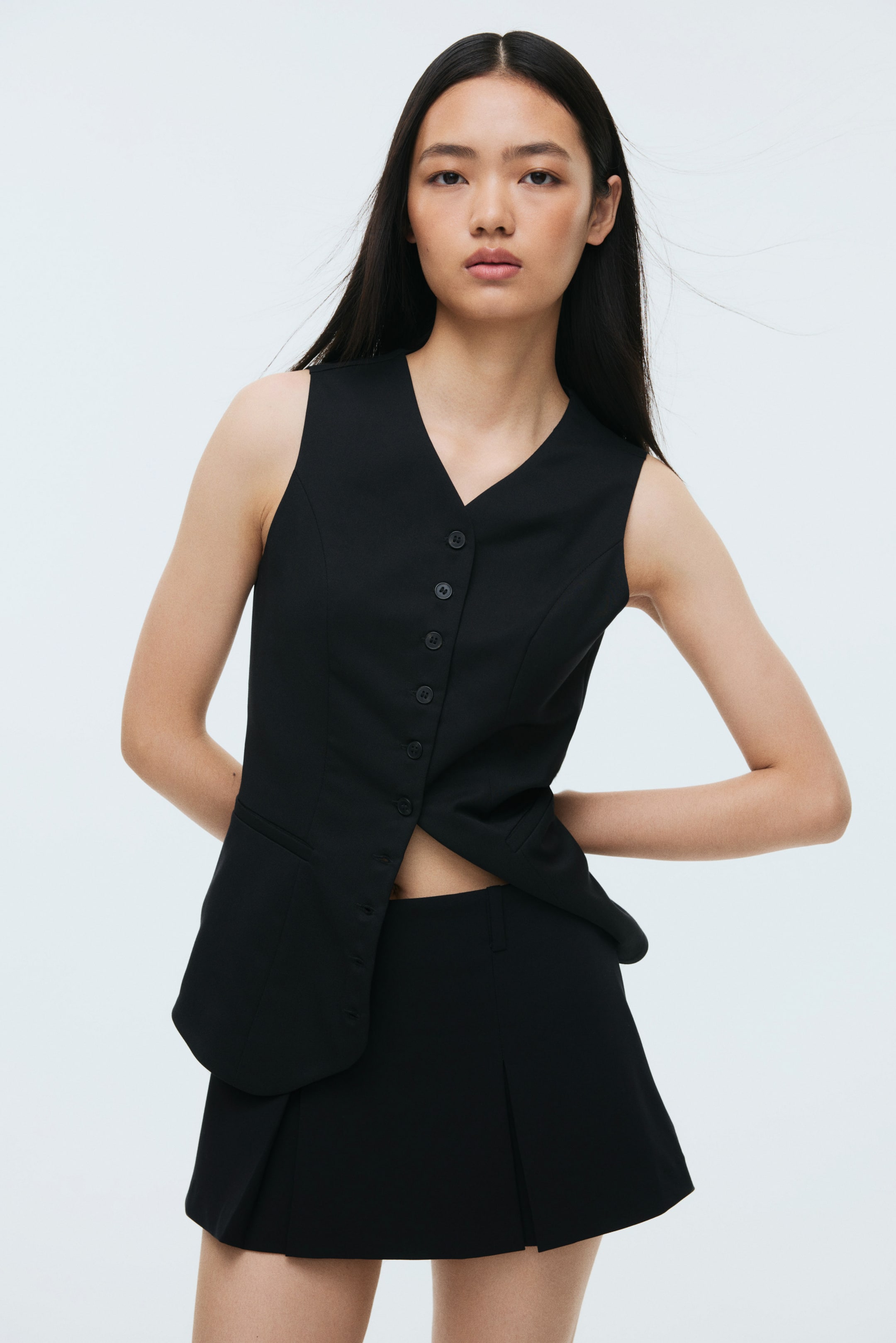 Ampliar la imagen: Un chaleco negro sin mangas, visible desde el frente, presenta un escote en V y seis botones frontales, con los más bajos desabrochados. Presenta dos bolsillos falsos de ojal y se combina con una minifalda negra, que muestra pliegues en su lado derecho.