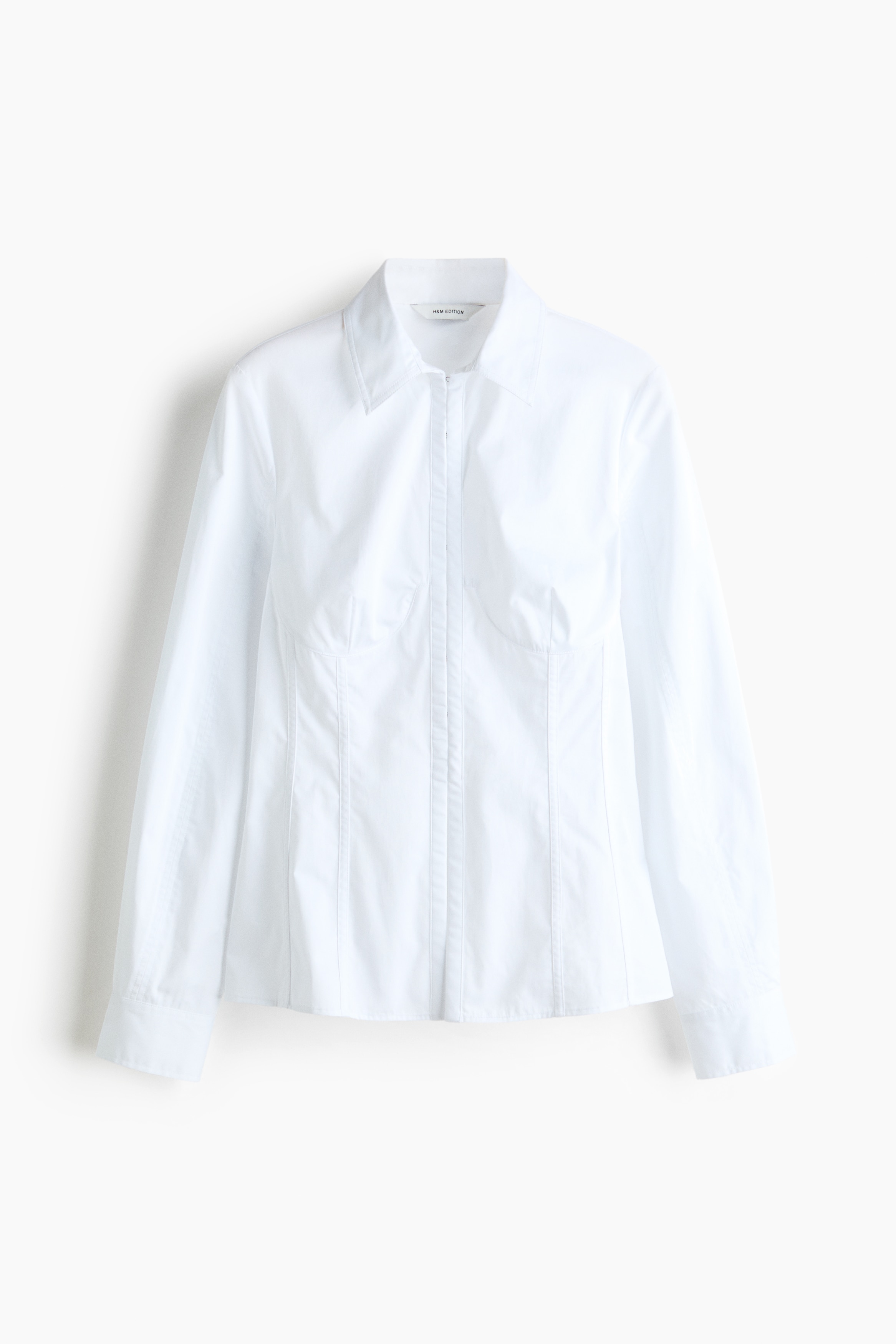 Una camicia bianca in cotone, dal taglio aderente, con colletto classico e maniche lunghe, Ã¨ indossata dalla modella. La camicia Ã¨ abbinata a una cintura corsetto nera ampia e strutturata, stretta in vita, e a pantaloni neri ampi a gamba larga dal taglio fluido.