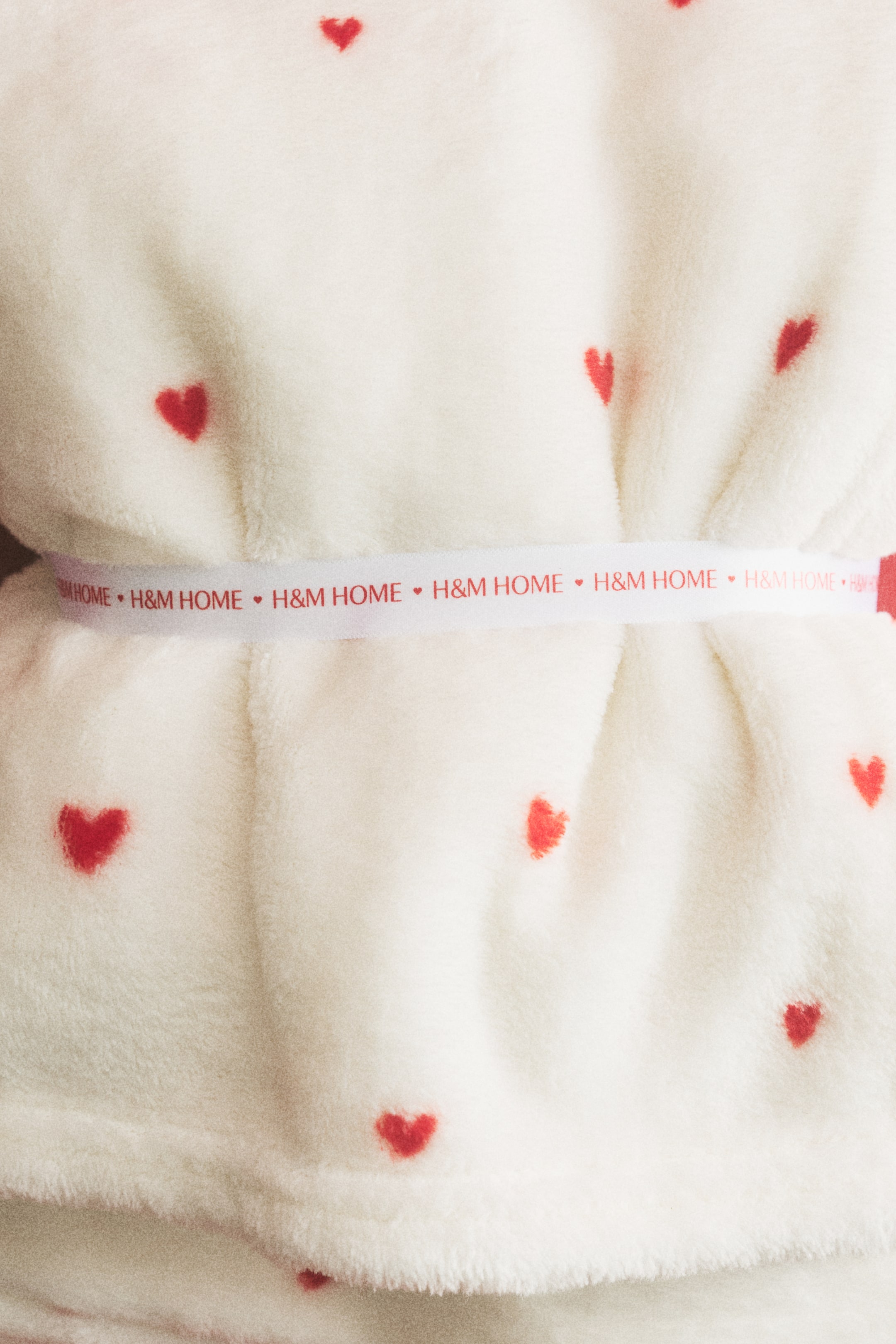 View larger image: A cream-coloured fluffy blanket patterned with red hearts is being presented, secured with a white ribbon bearing the repeating text 'H&M HOME' and small red hearts.