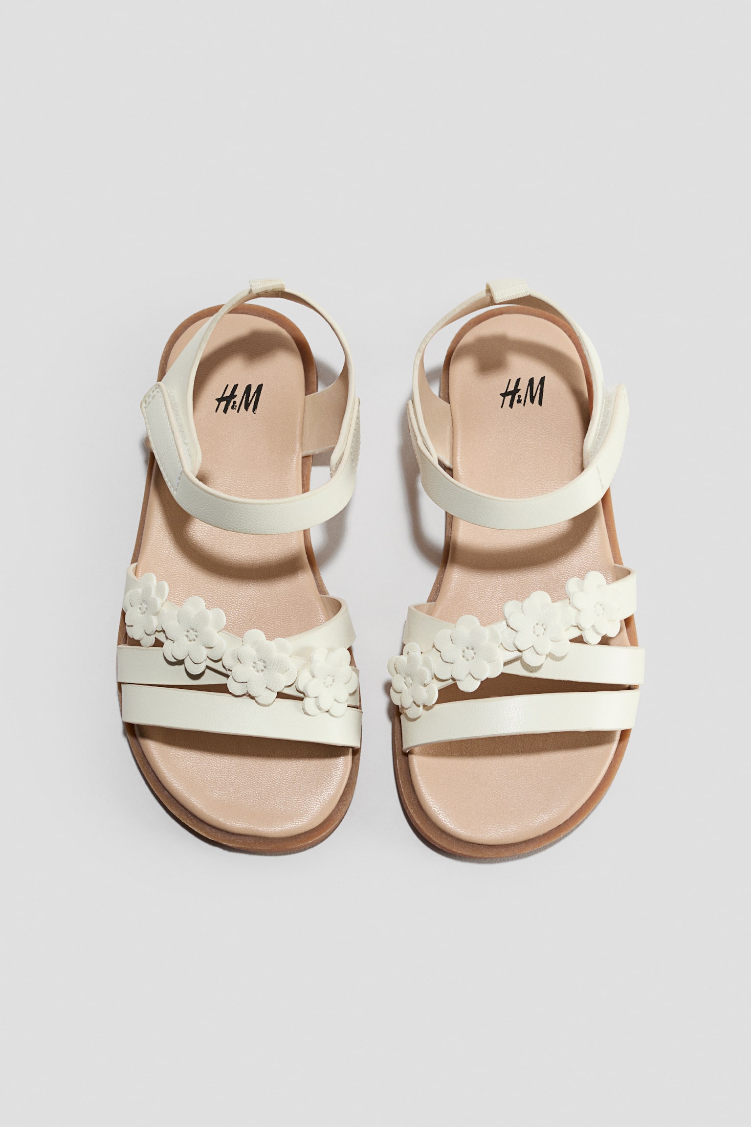 A pair of off-white children's sandals is facing front in a top-down view, featuring three narrow front straps with small flower appliquï¿½s embellishing the middle strap. The sandals are incorporating an adjustable ankle strap with a hook-and-loop closure and a light tan insole.