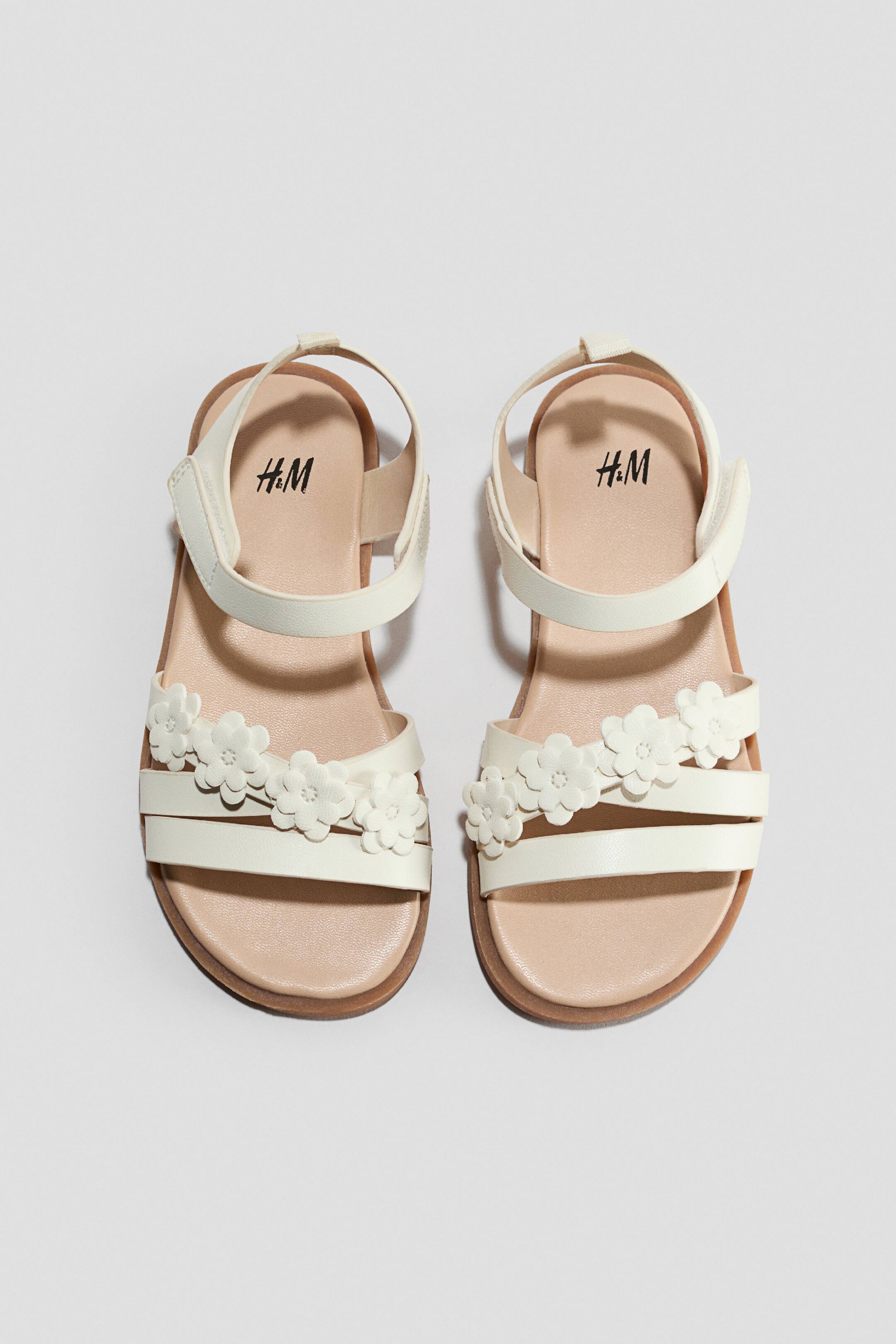 View larger image: A pair of off-white children's sandals is shown from a top-down front view, featuring three narrow front straps with small flower appliqués decorating the middle strap. The sandals include an adjustable ankle strap with a hook-and-loop closure and a light tan insole.