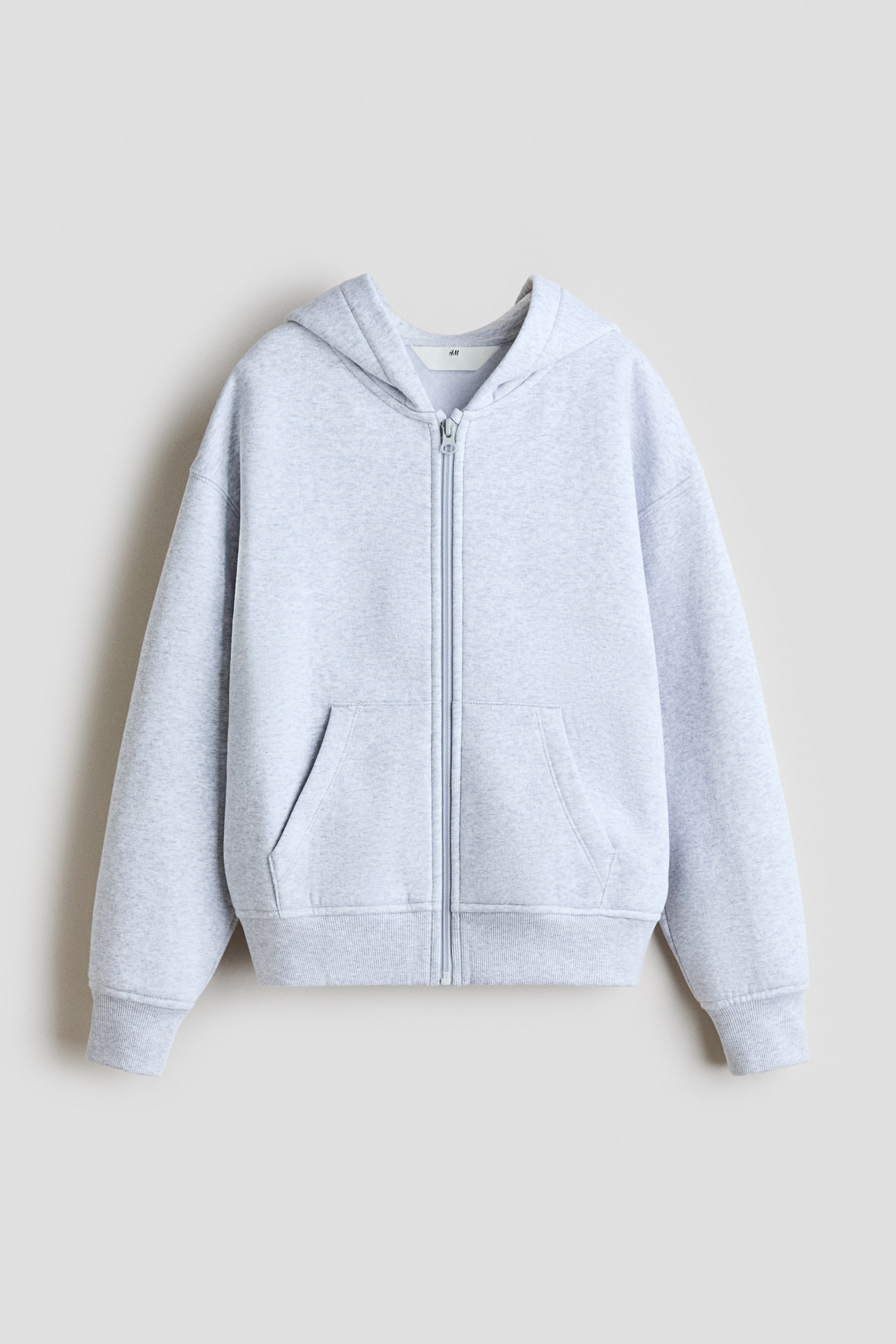 Visa större bild: En ljusgrå huvtröja i sweatshirtmaterial visas framifrån. Den har en dragkedja i full längd, en huva med jerseyfoder, nedhasade axlar och långa ärmar. Två framfickor, ribbade muddar och en ribbad nederkant är också synliga.