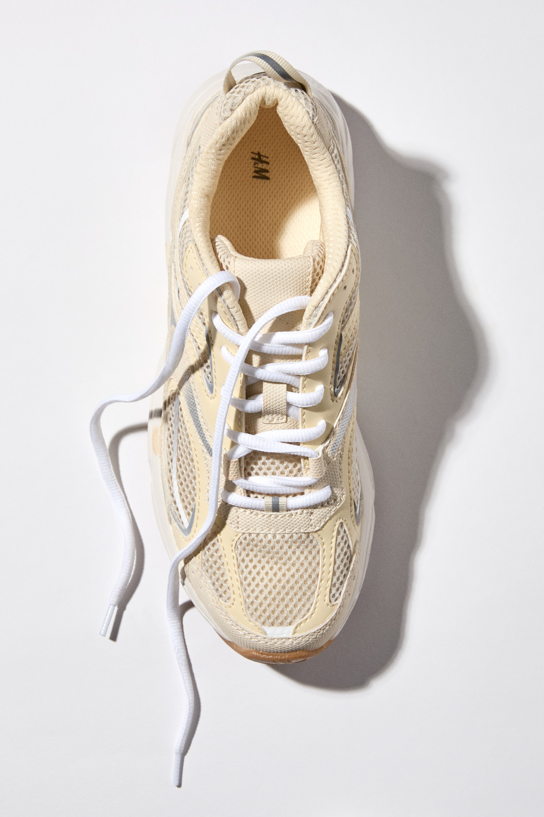 Größeres Bild anzeigen: Ein hellbeiger Sneaker wird aus einer Draufsicht präsentiert, mit Mesh-Einsätzen, synthetischen Overlays und silberfarbenen Akzenten. Weiße Schnürsenkel liegen ungebunden über dem Obermaterial, und ein H.M-Logo ist auf der Innensohle sichtbar.