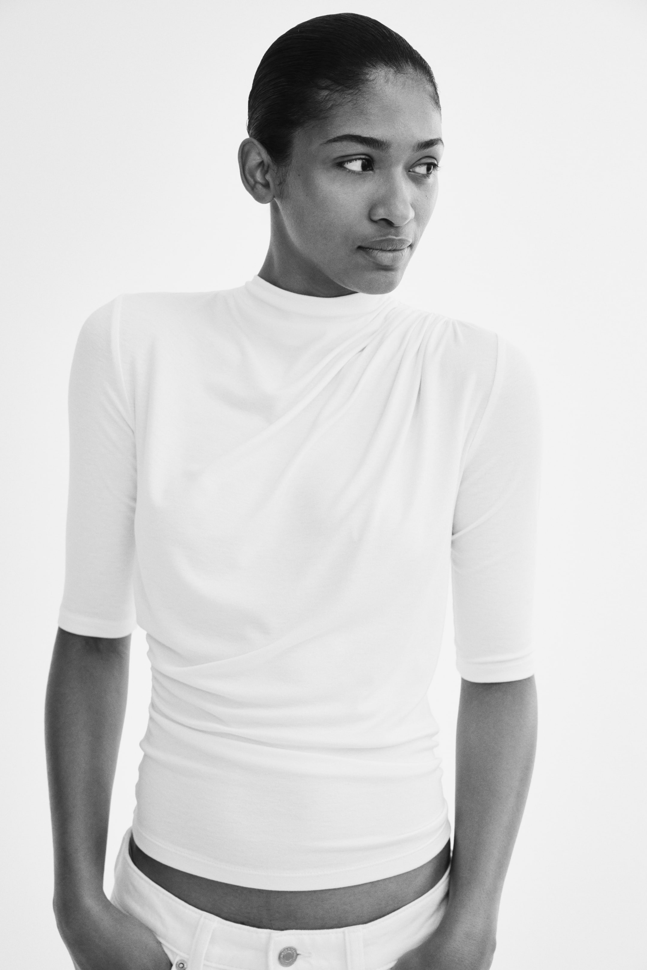 Ampliar la imagen: La modelo lleva una blusa blanca ajustada con cuello alto, mangas a la altura del codo y un detalle fruncido que crea un efecto drapeado en la parte delantera. La blusa se combina con pantalones blancos de tiro bajo.