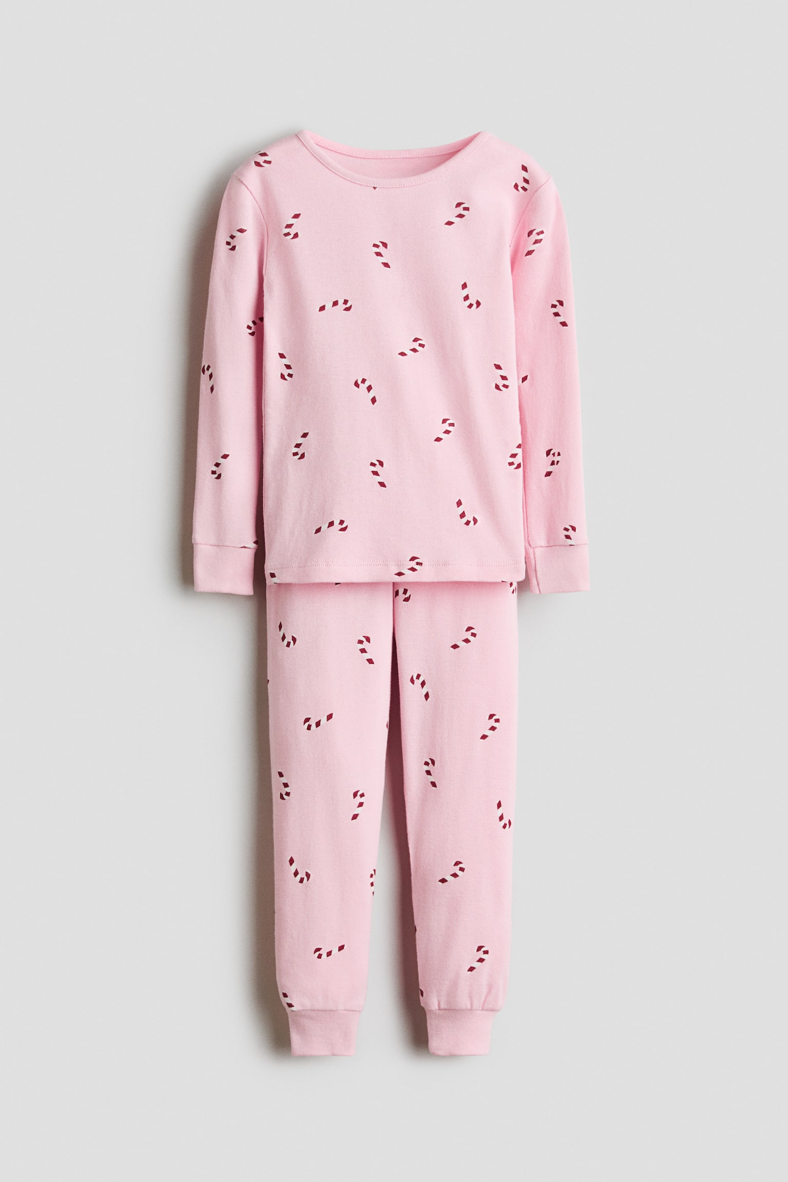 A front-facing view is showcasing a light pink cotton pyjama set featuring an all-over red candy cane print. The set is displaying a long-sleeved top with a round neckline and ribbed cuffs, paired with matching pants that are tapering into ribbed hems.