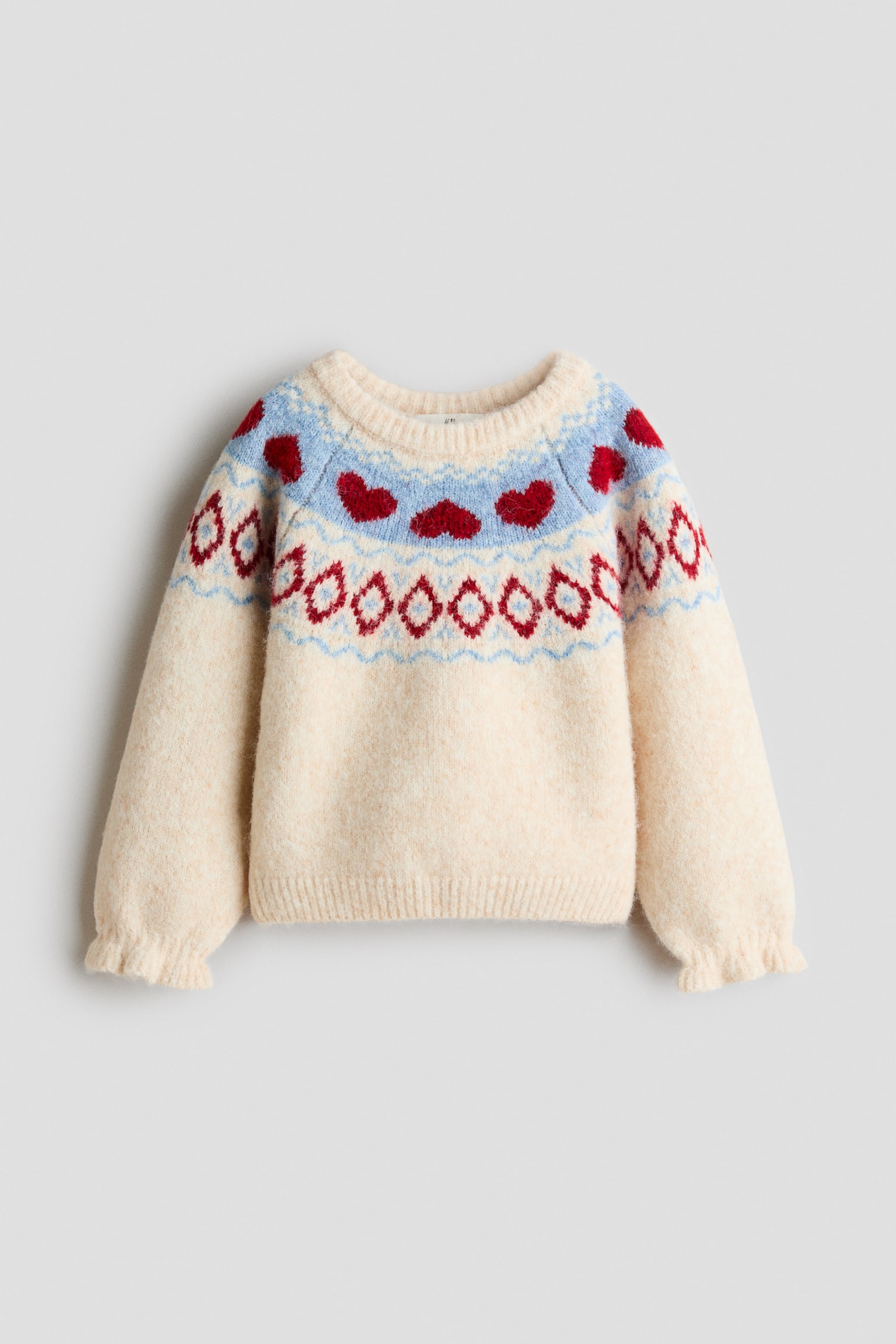 A cream-colored knit sweater is facing front, showcasing a decorative circular yoke pattern with red hearts on a light blue band and red geometric diamond shapes. The sweater is featuring a ribbed crew neck, a ribbed hem, and long sleeves with gathered, ruffled cuffs. - 1