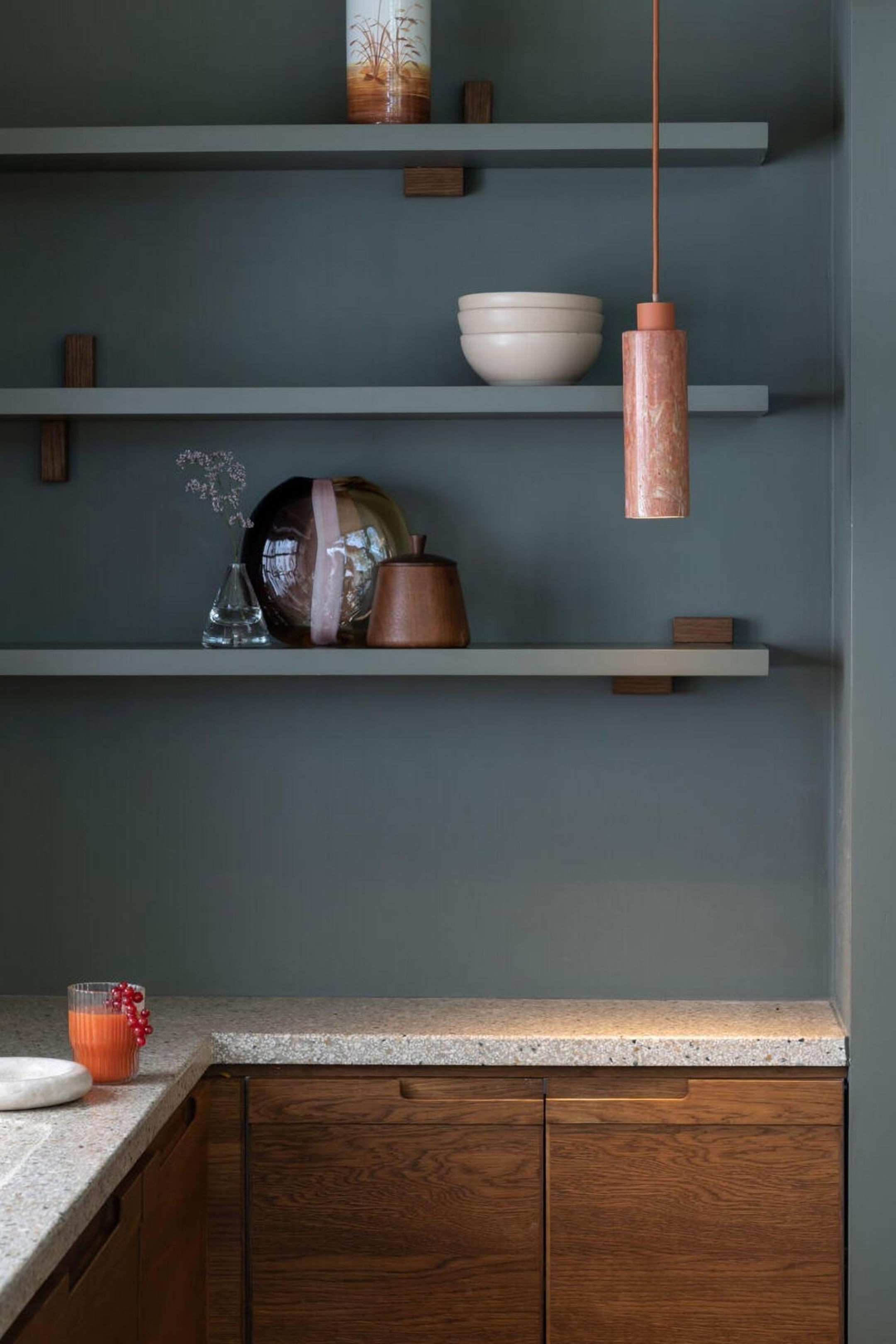 Grotere afbeelding bekijken: Een cilindervormige hanglamp van roze-oranje gemarmerde steen hangt aan een slanke koord en is voorzien van een bijpassende terracotta kleurige kap aan de bovenkant, terwijl ze een zachte neerwaartse gloed uitstraalt.