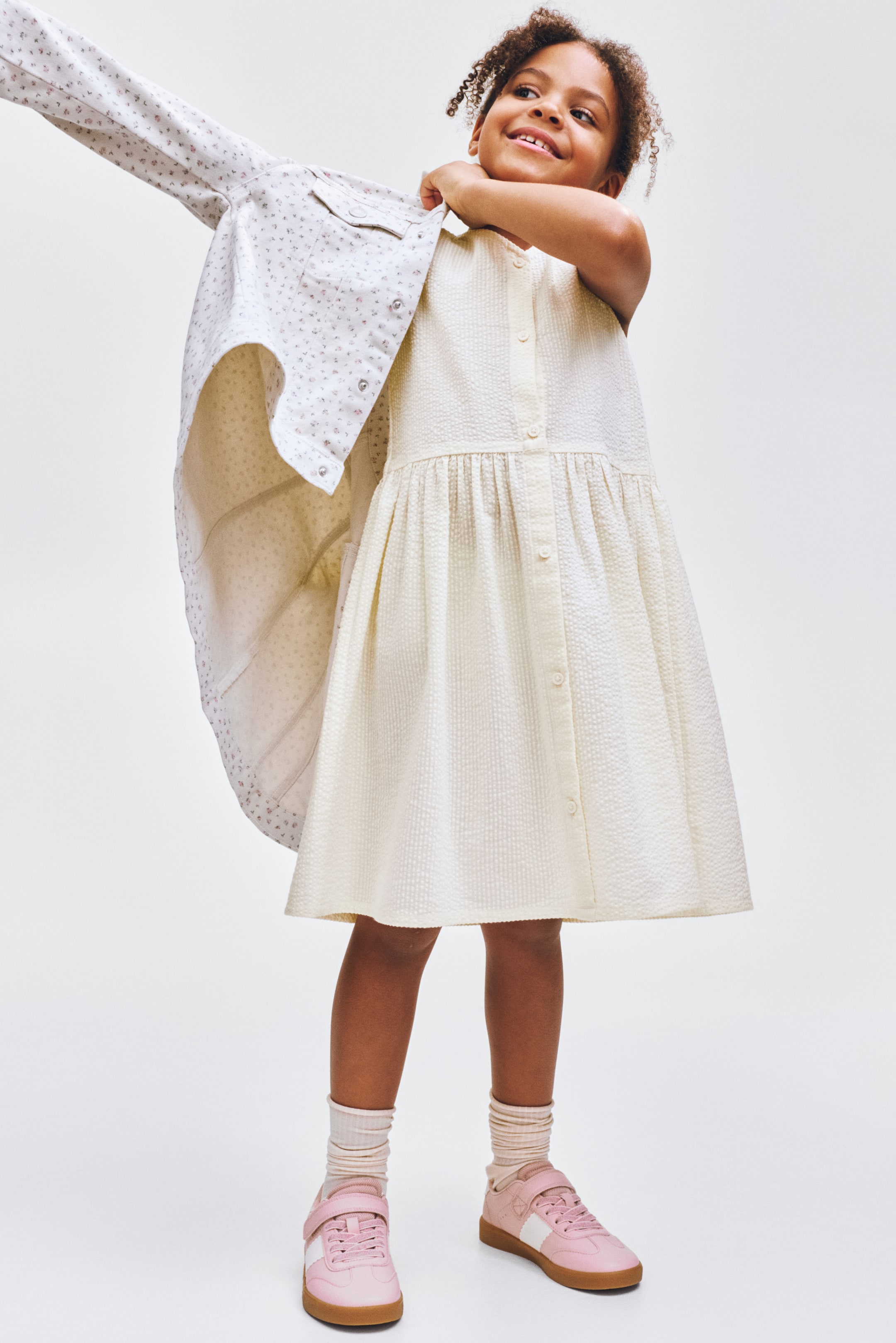 Xem ảnh lớn hơn: Một chiếc váy không tay màu kem với kết cấu dệt, phần thân trước có nút bấm và phần eo được thu gọn chảy vào chân váy xòe đang được mặc, trong khi một chiếc áo khoác trắng với họa tiết chấm nhỏ màu hồng và nút bấm đang được khoác lên trên.