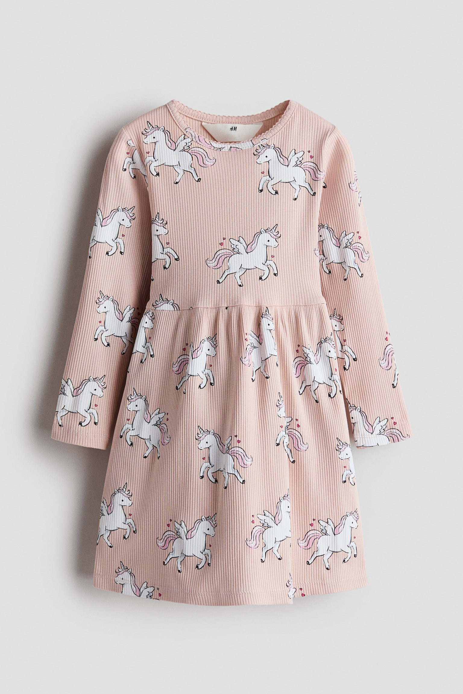 A front view of a pink long-sleeved dress in ribbed knit fabric, featuring an all-over pattern of white winged unicorns with pink manes and tails. The dress is designed with a round neckline trimmed with picot, a gathered seam at the waist, and a flared skirt.