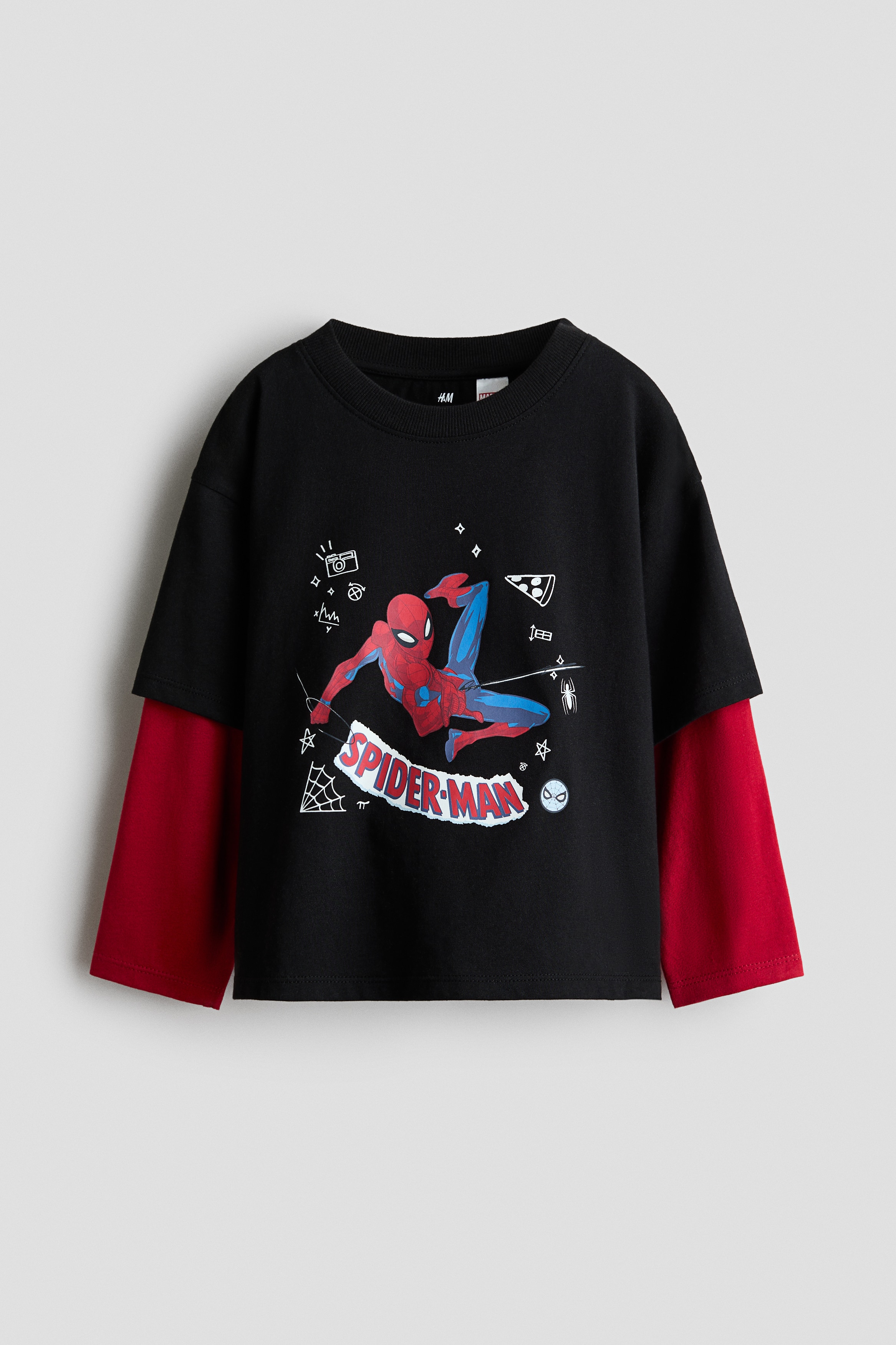 A black cotton jersey tee featuring a printed Spider-Man graphic on the front is worn by the model. The tee features a layered design with dropped shoulders, short black sleeves, a ribbed crew neck, and sewn-in long red sleeves, styled with black cargo trousers.