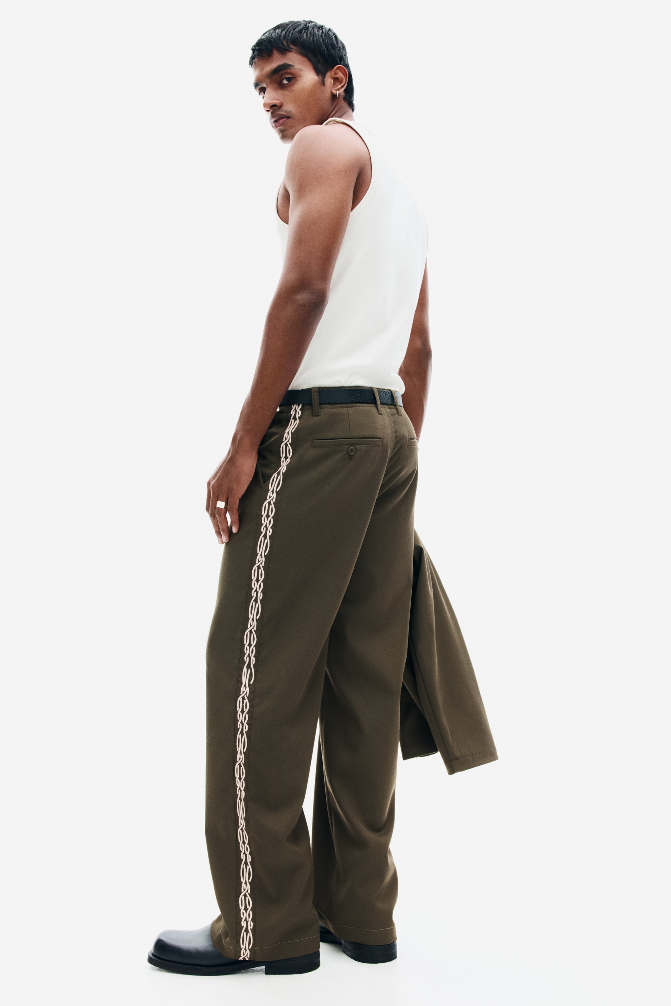 View larger image: The model is wearing olive-green relaxed-fit chinos featuring a cream-coloured embroidered chain pattern running down the side of the leg. The trousers are styled with a black belt and a white tank top, while a matching olive-green jacket is being held in one hand.