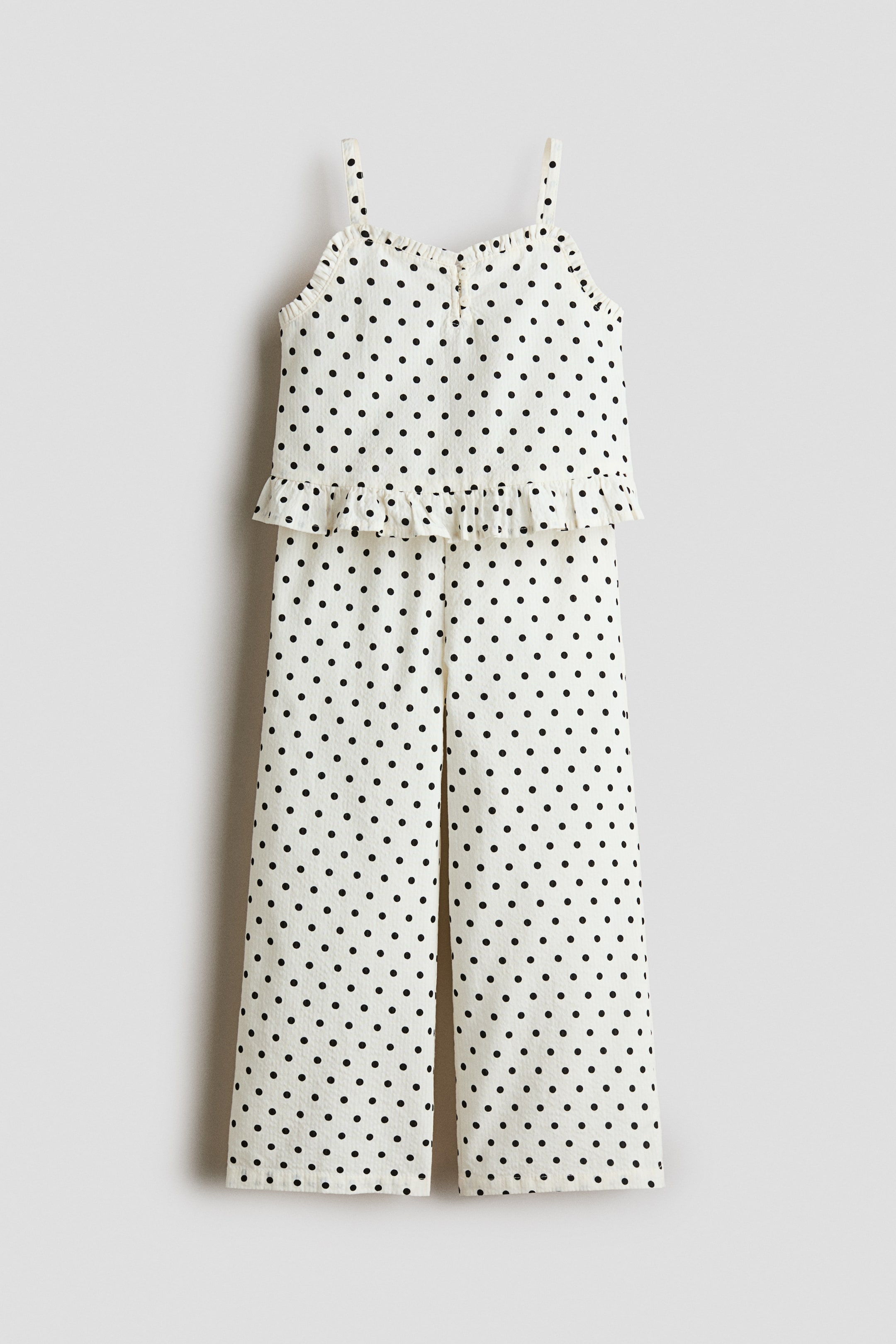 View larger image: A cream-coloured jumpsuit with a black polka dot pattern is shown from the front, featuring thin shoulder straps and a ruffled trim along the neckline. It includes a ruffled peplum-style overlay at the waist and falls into wide-leg trousers.