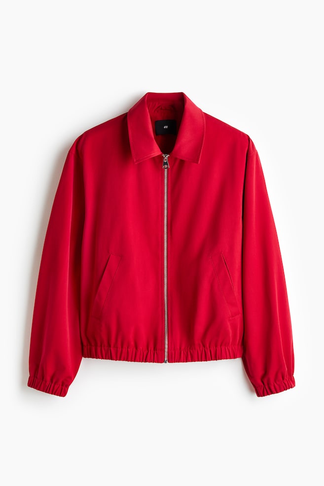 A red jacket is facing front, featuring a turn-down collar, a zipper running down the front, two front welt pockets, and covered elastication at the cuffs and hem. - 1