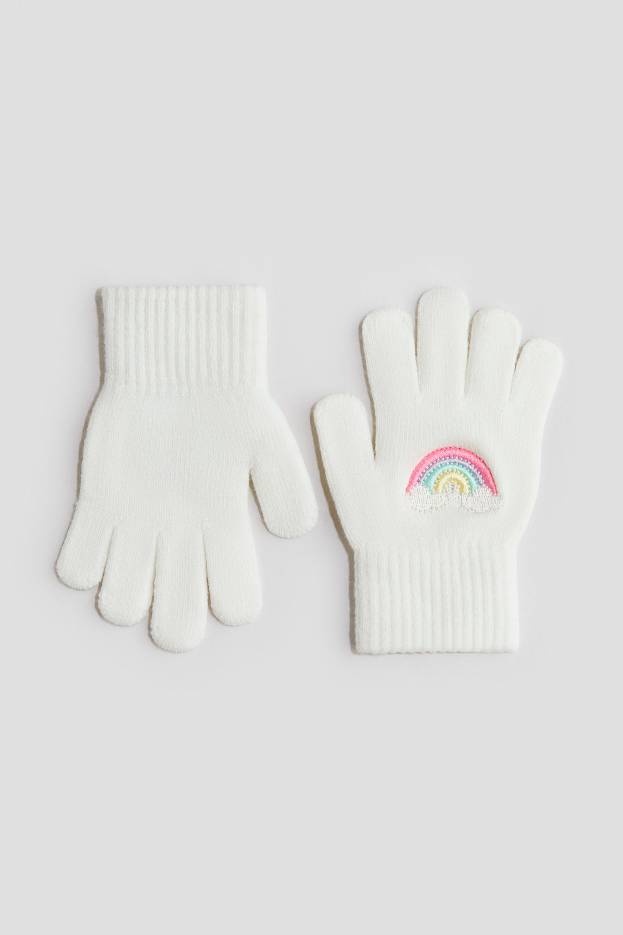 Voir une image plus grande: Une paire de gants tricotés blancs avec poignets côtelés. Un gant montre sa paume unie, tandis que l’autre affiche au dos un motif arc-en-ciel brodé et coloré avec un nuage blanc.
