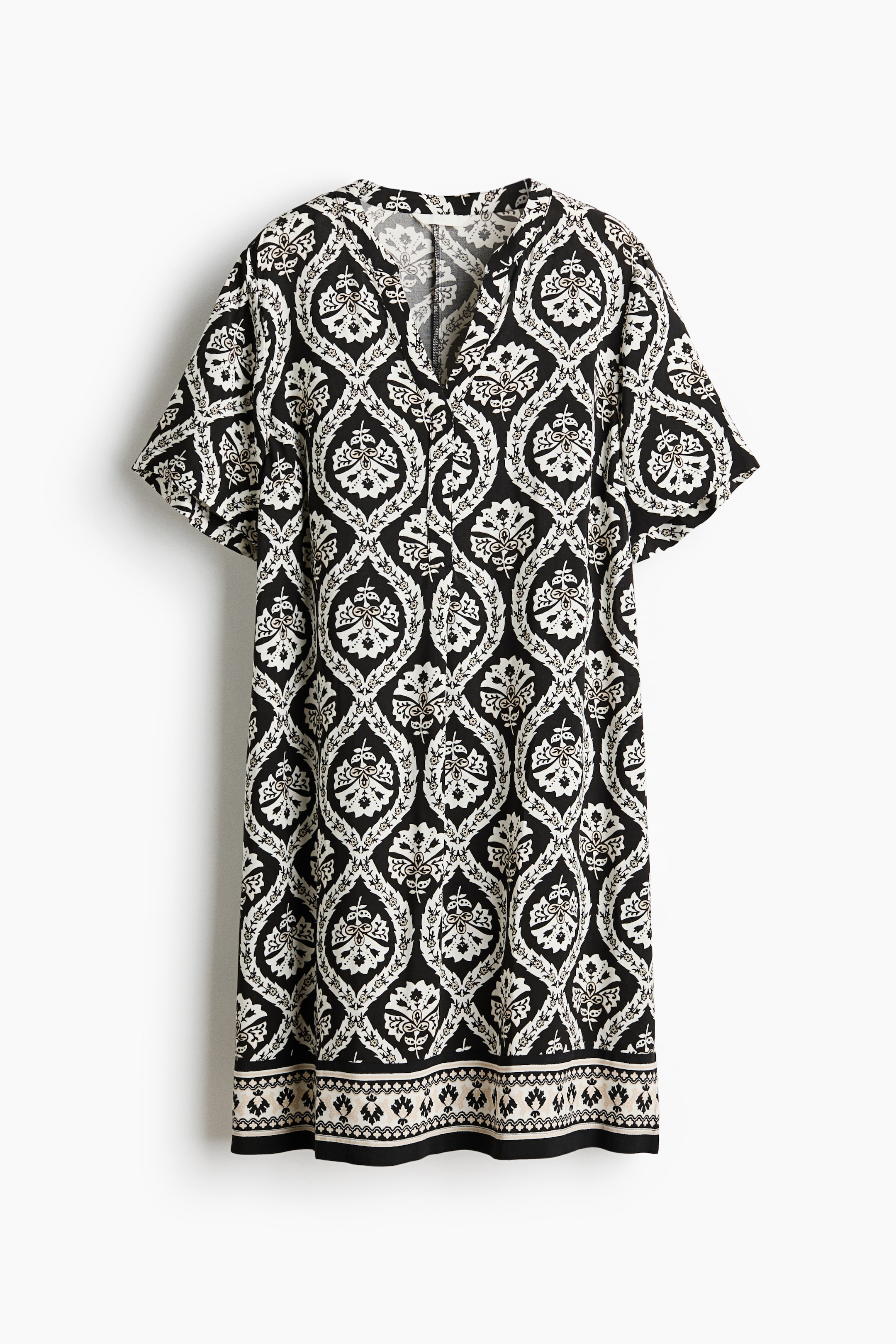 Agrandir l'image: Une robe tunique longueur genou est présentée de face, affichant un motif ornemental noir et blanc. Elle est dotée d'un col officier, d'une ouverture en V, de manches courtes avec des épaules légèrement tombantes, et d'un ourlet droit avec une bordure à motif distinctif.