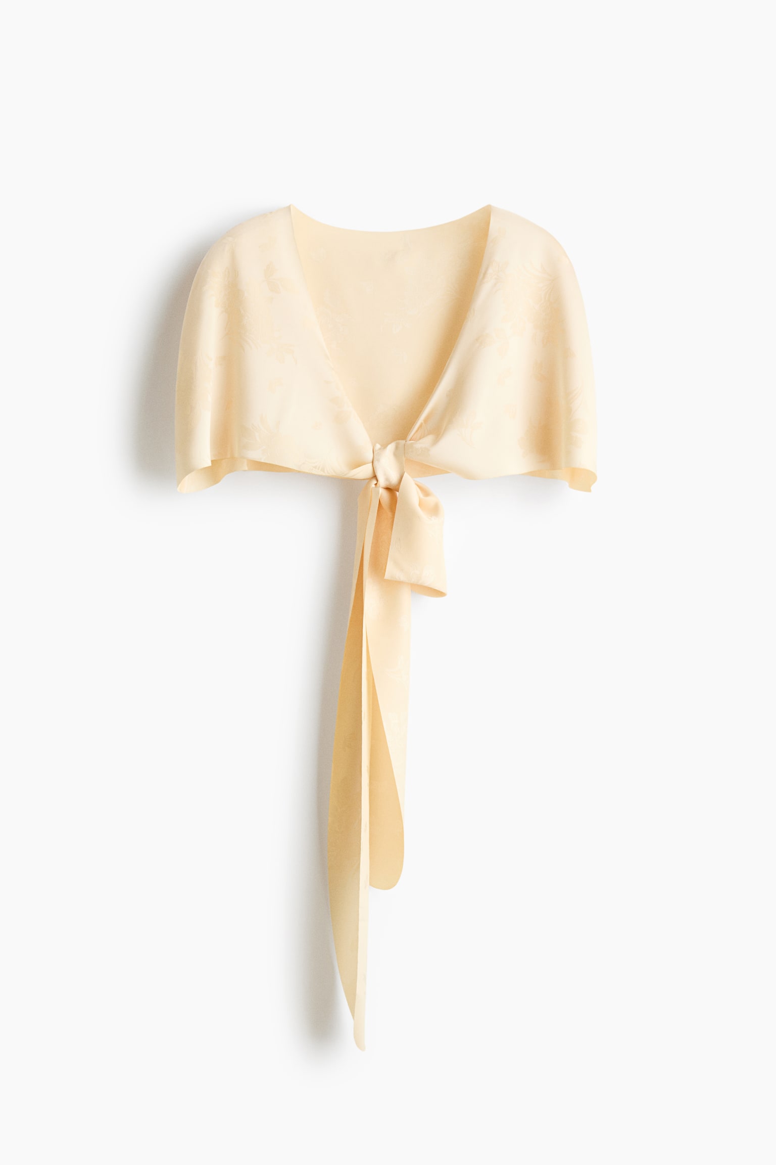 A pale yellow jacquard capelet is shown from the front, featuring short draped sleeves and a subtle floral pattern. The garment fastens at the centre front with a knotted tie that creates long, hanging fabric tails.