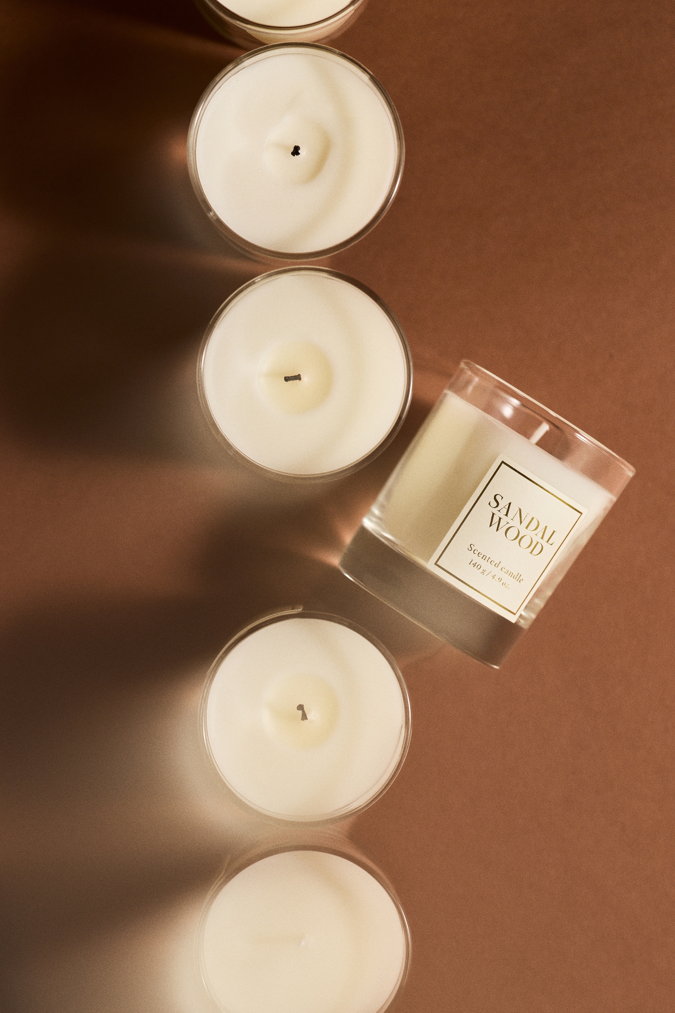 A scented candle in a clear glass holder with a thick base is standing next to its brown, patterned gift box. Both the candle and the box are shown from a front-facing angle, featuring matching square white labels with gold borders and the text "SANDAL WOOD".