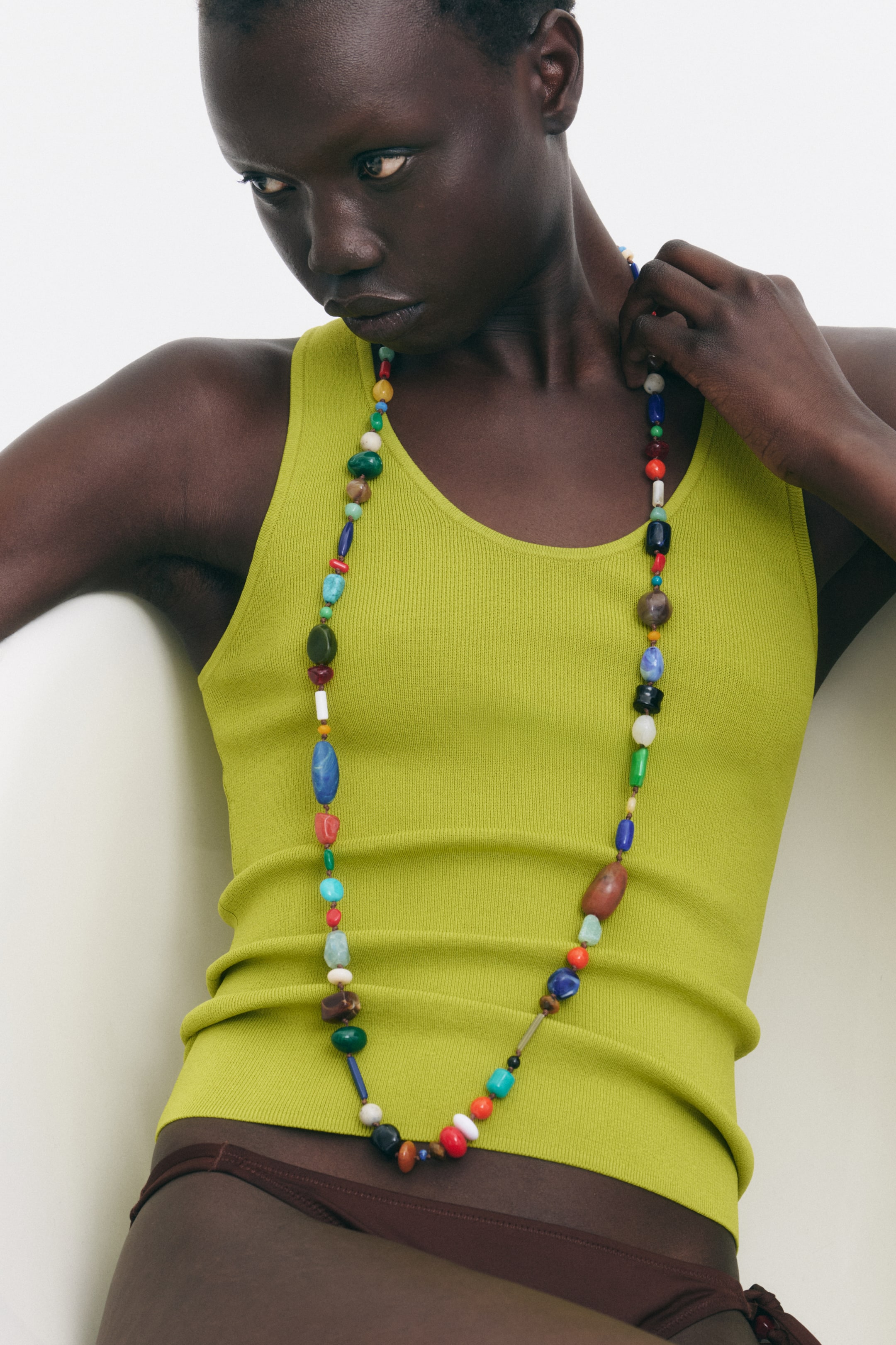 View larger image: The model is showcasing a long, knotted cord necklace adorned with plastic beads in an array of shapes and colours. She is seen adjusting the necklace at the nape of her neck.