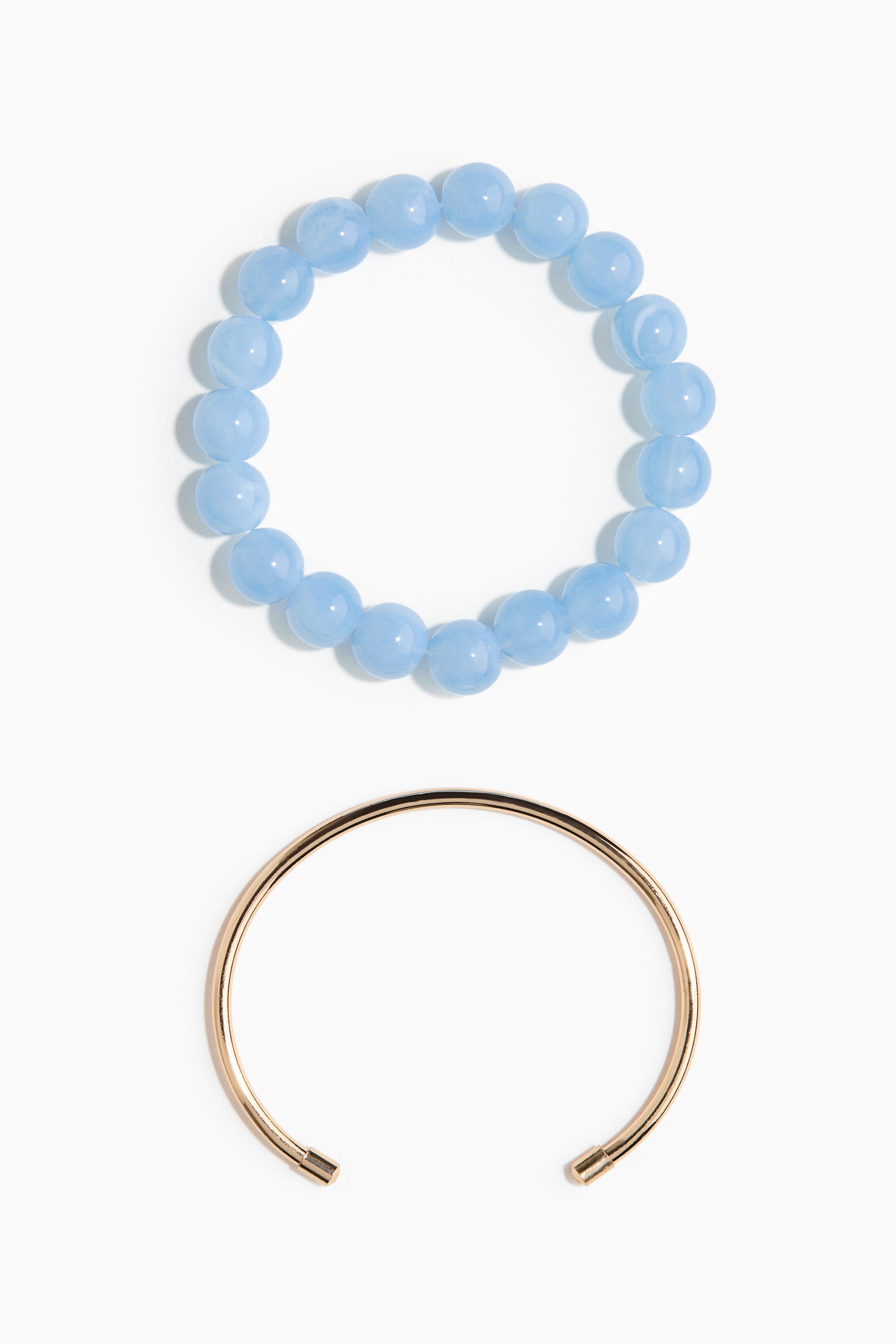 View larger image: The top bracelet is a stretch design featuring round, light blue, translucent beads, while the bottom bracelet is a smooth, gold-toned open cuff with cylindrical caps on its ends.