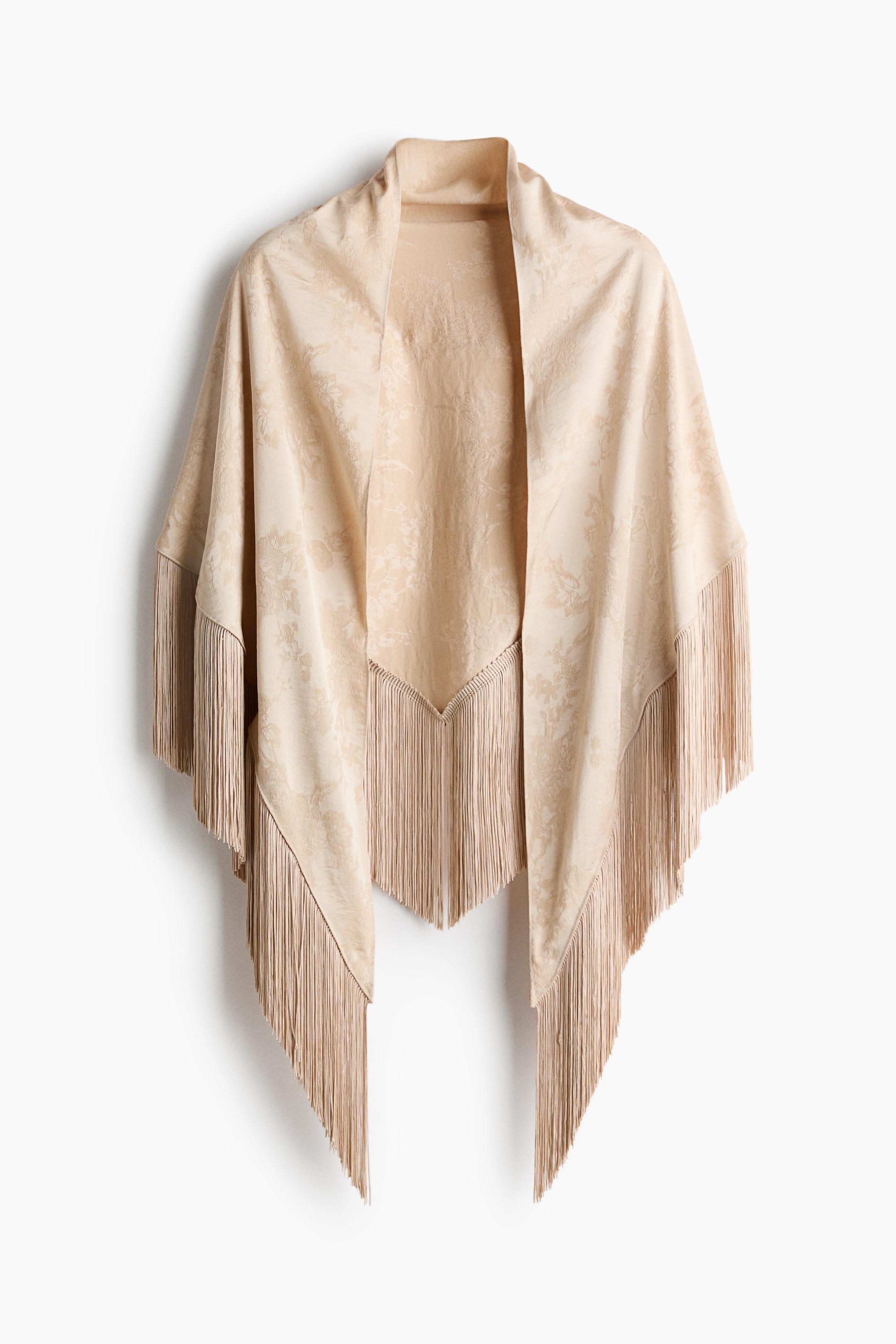 View larger image: A beige triangular scarf in a floral jacquard weave with a subtle sheen is draped forward in a front view, showcasing how it falls over the shoulders. The design features long, matching fringes hanging straight down along the scarf’s edges.