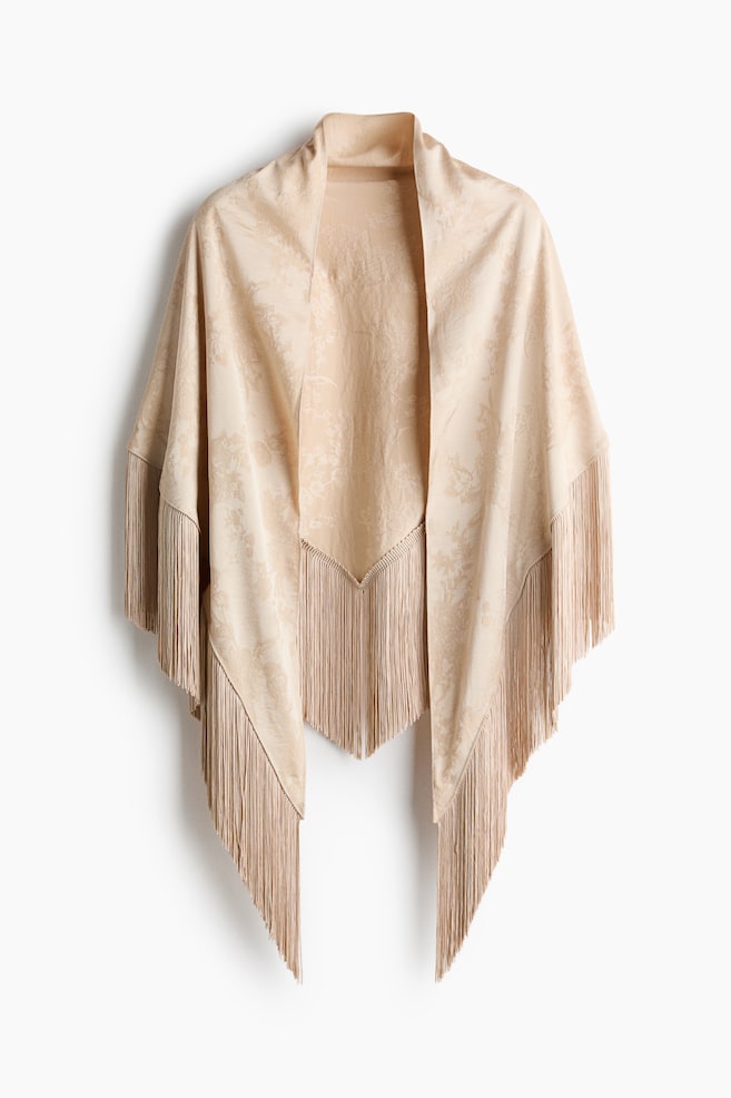 Et beige trekantsjal i en floral jacquardvævning med en diskret glans hænger fremad set forfra og viser, hvordan det falder over skuldrene. Designet har lange, matchende frynser, der hænger lige ned langs tørklædets kanter. - 1