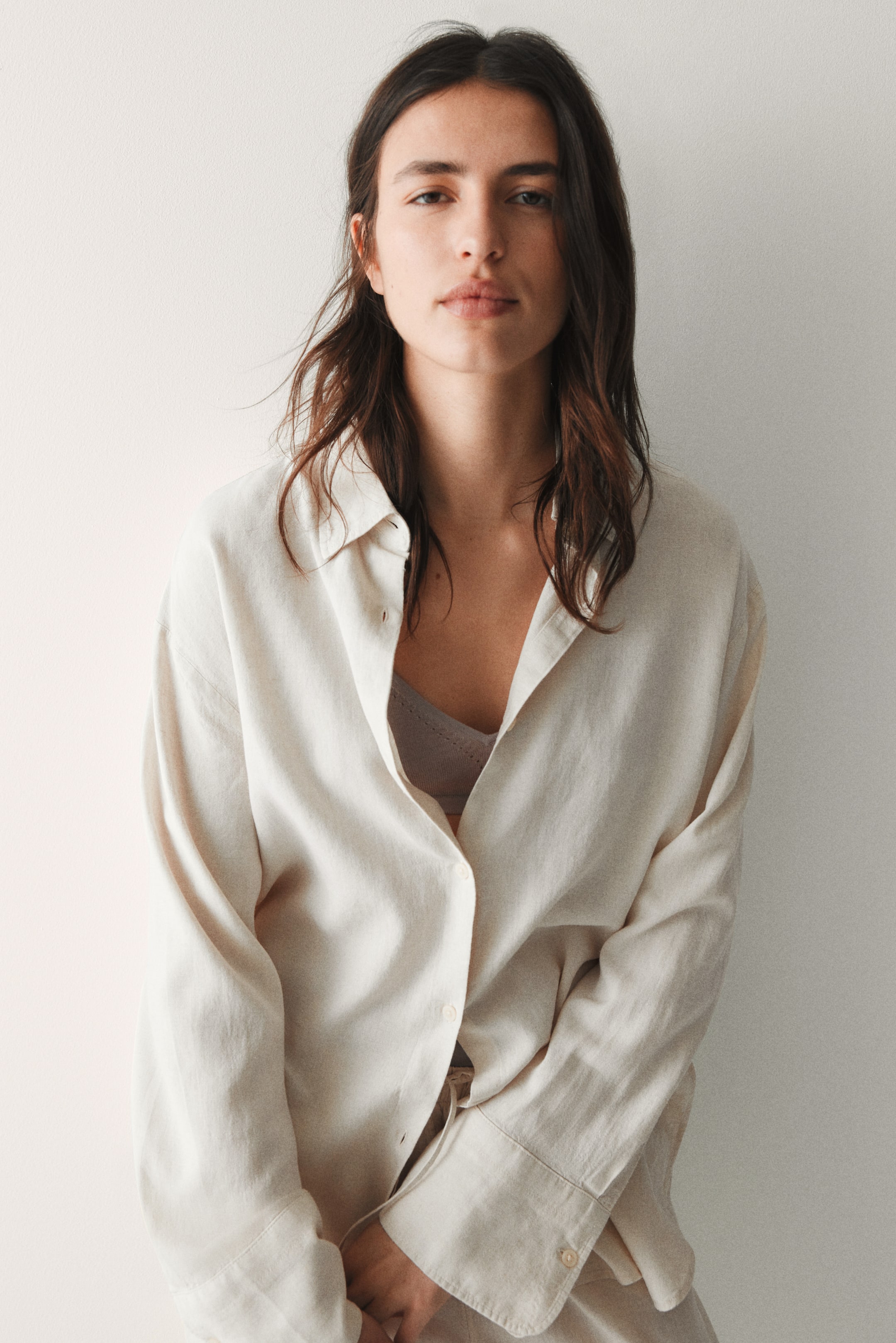 View larger image: An oversized, light-coloured button-up shirt is styled from a front-facing perspective, left unbuttoned to reveal a grey bralette beneath. The shirt features a pointed collar, prominent front buttons, and long sleeves with unfastened cuffs, all enhancing its laid-back fit.