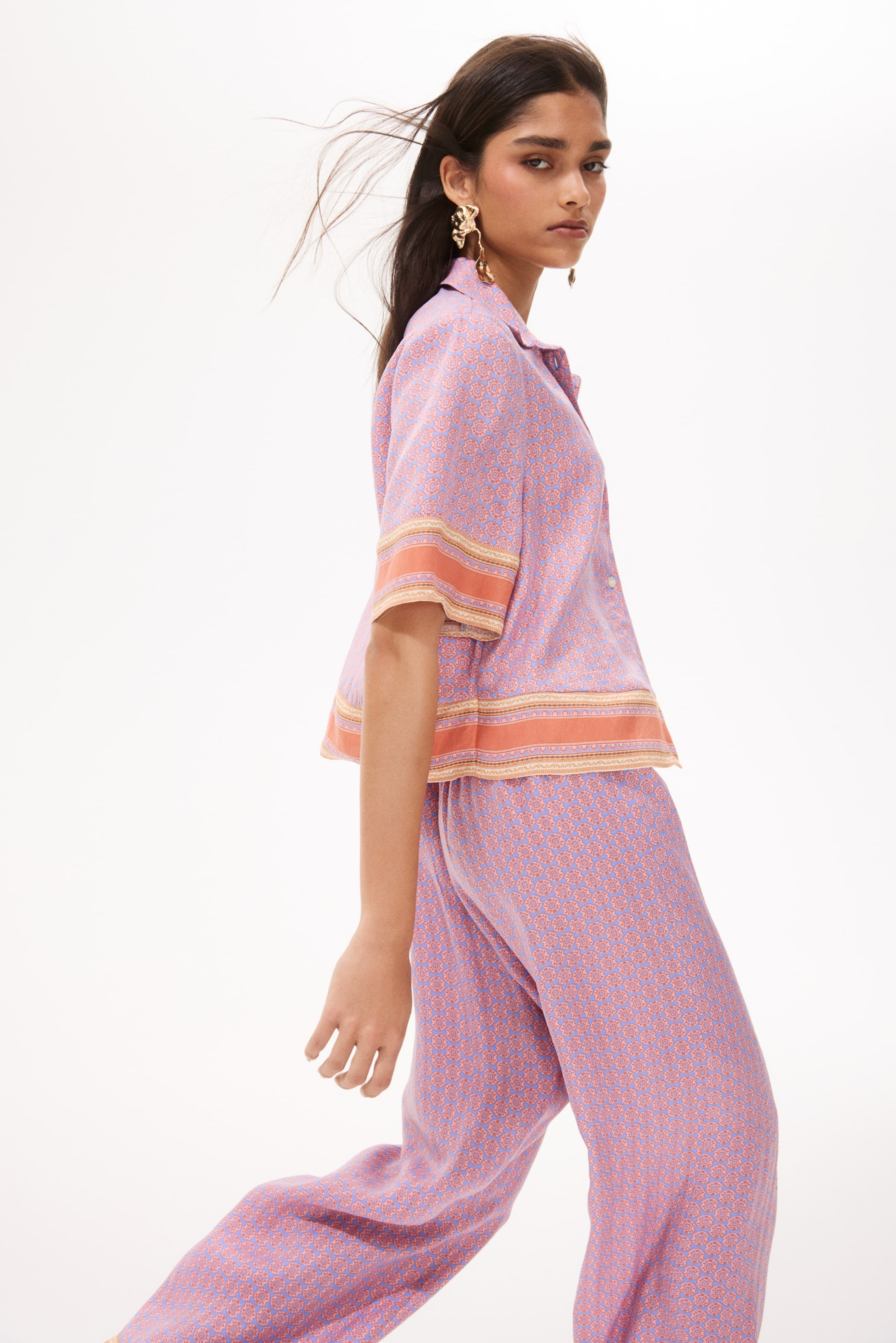 View larger image: A matching short-sleeved shirt and wide-leg pants set is displayed from a side-front angle. The ensemble showcases a light purple fabric adorned with a repeating orange geometric pattern. The shirt features short sleeves, a camp collar, and a distinctive orange and gold striped border on the sleeve cuffs and hem. Meanwhile, the wide-leg pants continue the same purple and orange geometric pattern.
