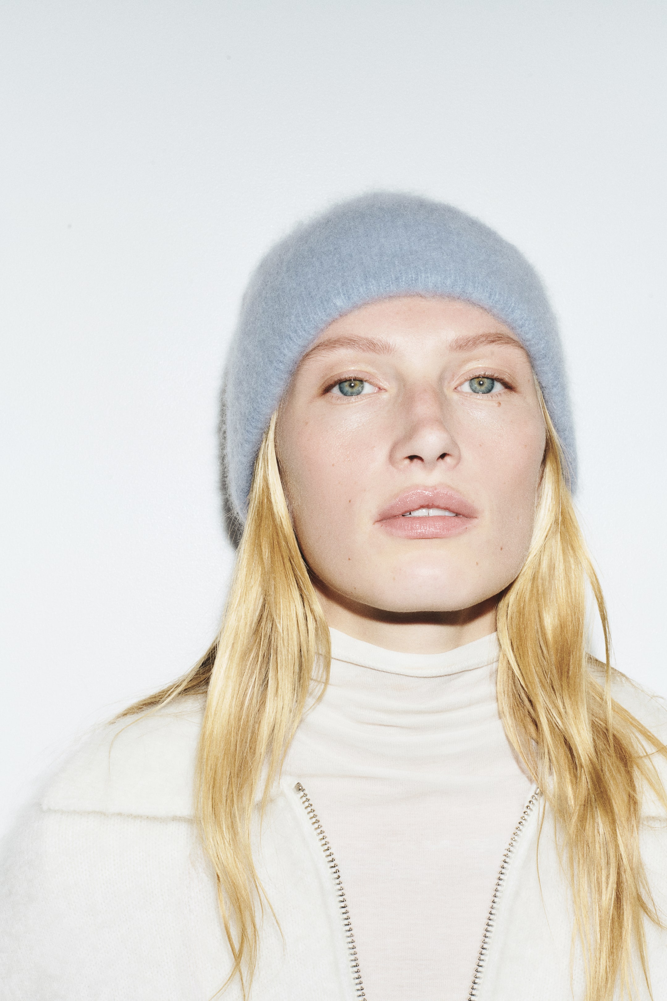 Преглед на по-голямо изображение: Светлосинa шапка тип beanie от мека, четкана плетка с подгънат ръб е носена на главата от модела. Шапката е стилизирана в комбинация с кремав топ с висока яка и бяло плетено яке с цип.