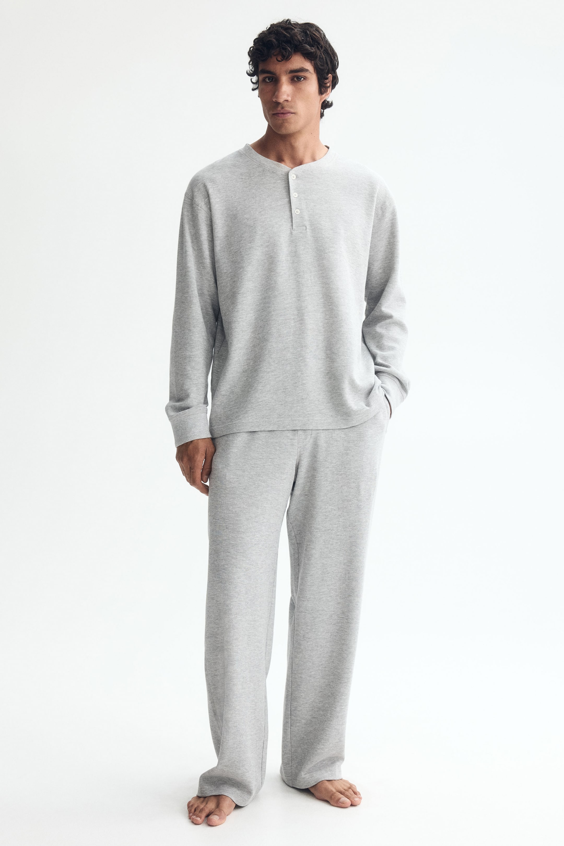 Agrandir l'image: Un ensemble de vêtements de détente gris, composé d'un t-shirt Henley à manches longues avec une patte de boutonnage à trois boutons et d'un pantalon de détente assorti à la coupe ample, est porté par le mannequin. Le mannequin se tient debout avec une main dans la poche du pantalon.
