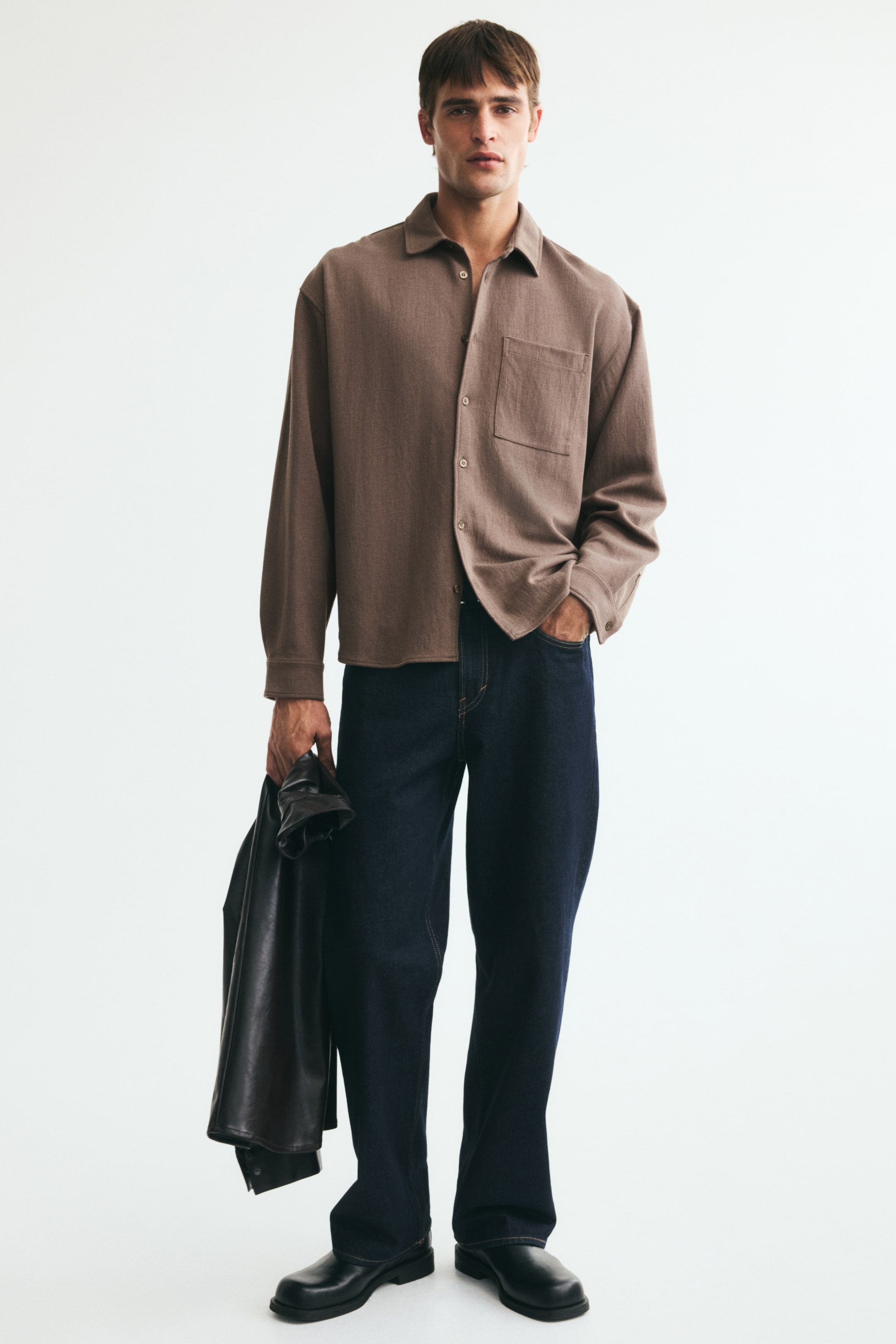 View larger image: A brown interlock jersey shirt with a collar, French front, and an open chest pocket is being worn by the model. The shirt features dropped shoulders, long sleeves with buttoned cuffs, a gently rounded hem, and a relaxed fit.