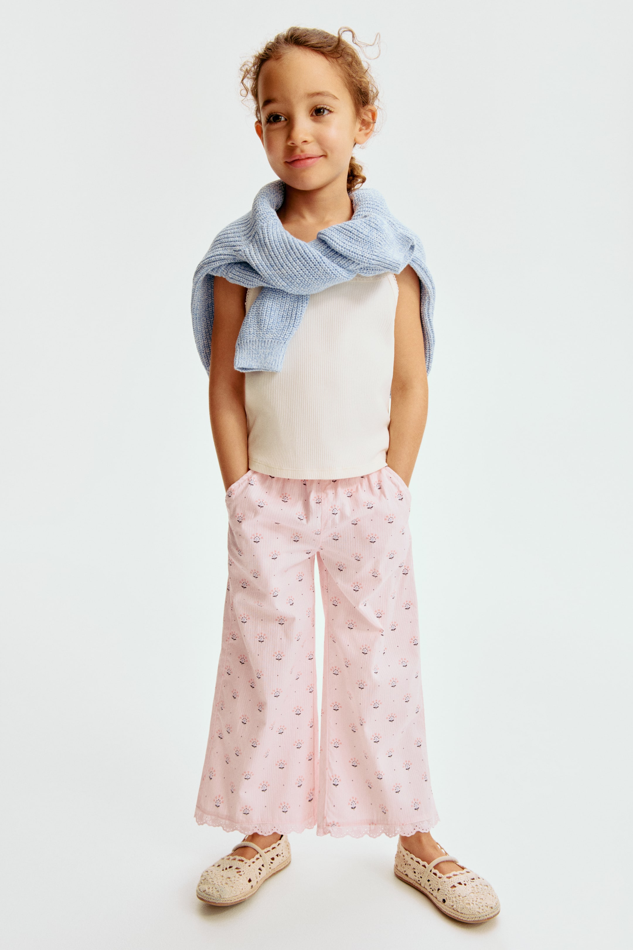 View larger image: A light blue ribbed knit sweater is layered over a white ribbed tank top. Pink wide-leg trousers showcase white pinstripes, a floral pattern, and a scalloped lace hem, paired with cream crochet espadrille flats.