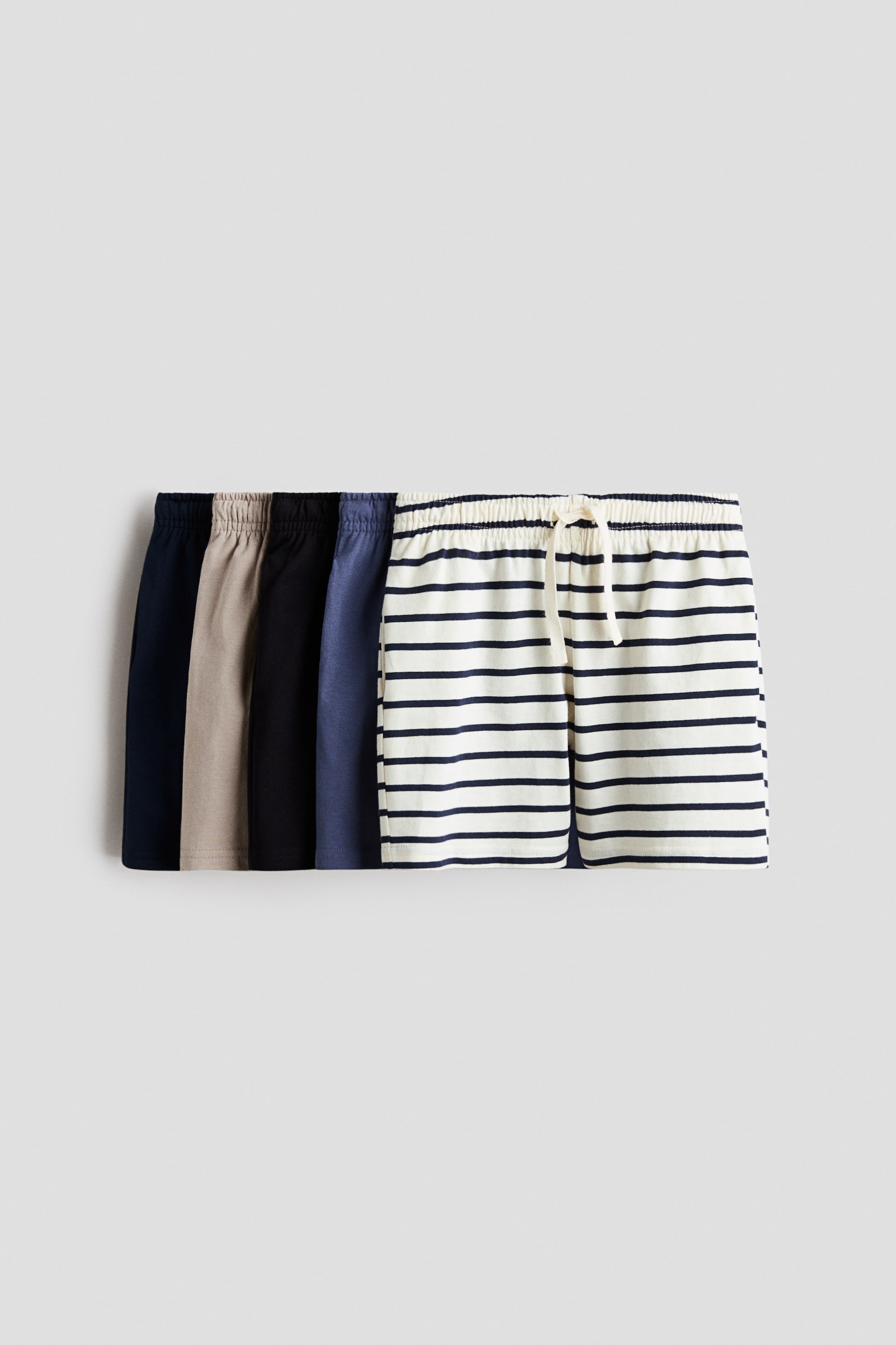 Visualizza immagine più grande: Sono esposte cinque paia di pantaloncini, ciascuno con vita elasticizzata con coulisse e in morbido jersey di cotone. Un paio, rivolto frontalmente, è bianco con righe orizzontali blu navy e una coulisse di colore chiaro, mentre altri quattro paia sono parzialmente visibili in tinta unita blu navy, beige, nero e blu scuro.
