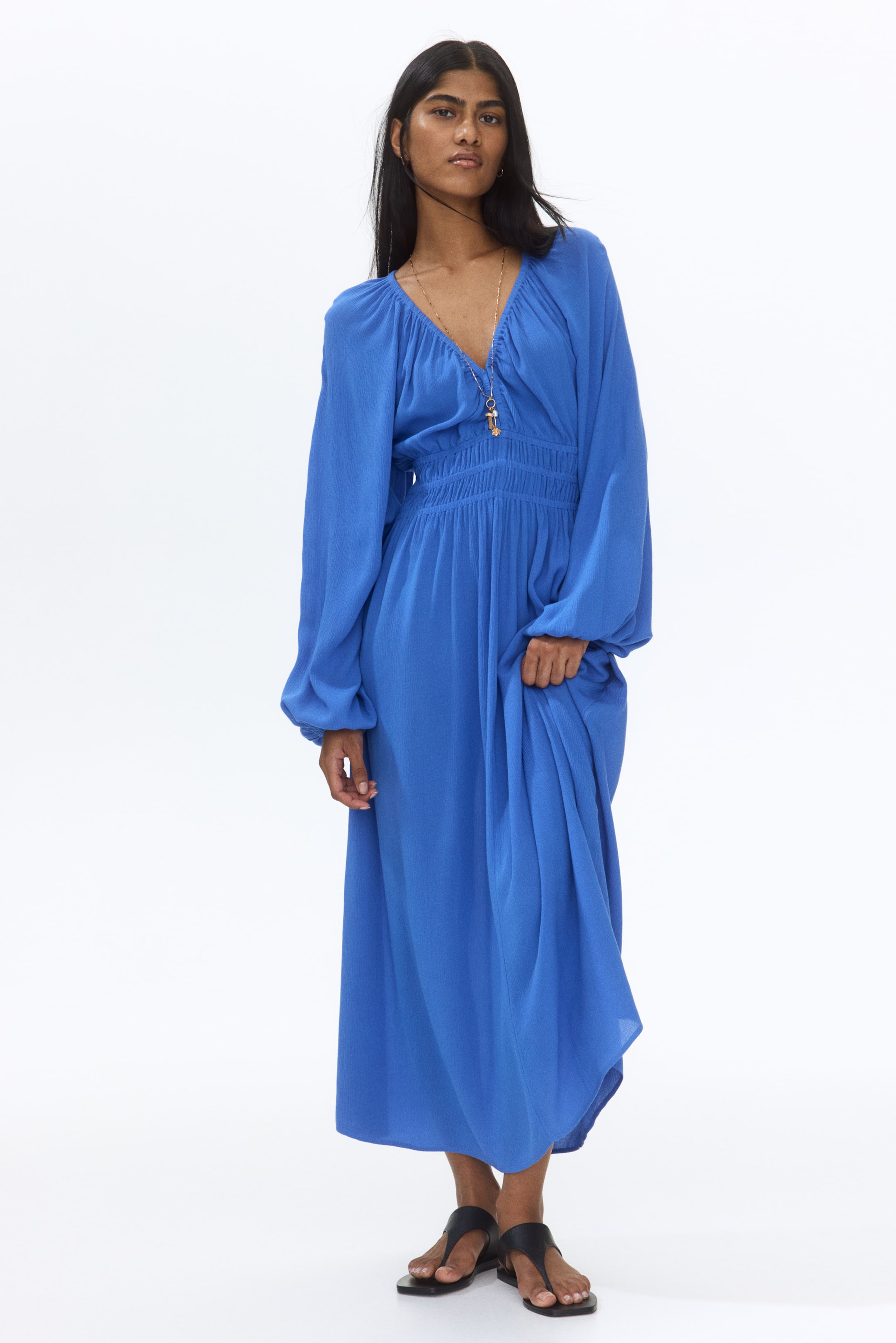View larger image: The blue maxi dress showcases a deep V-neckline, long balloon sleeves with elasticated cuffs, and a ruched waist. A long necklace with a pendant graces its V-neck, while a hand elegantly lifts a portion of the flowing skirt.
