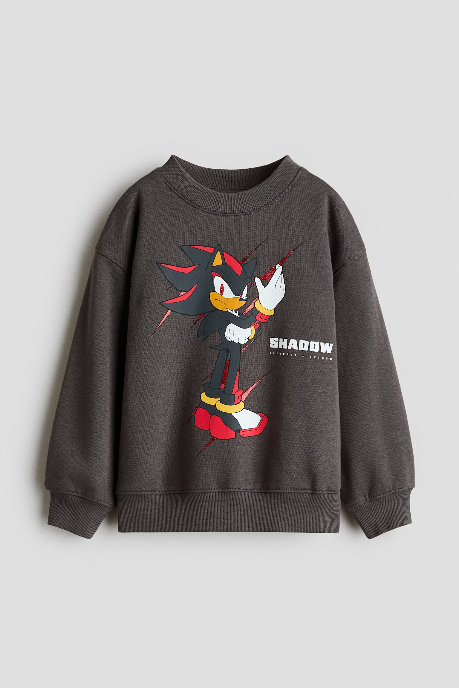Een donkergrijze, losvallende sweatshirt, van voren weergegeven, met een grafische print van het personage Shadow the Hedgehog en de tekst "SHADOW" op de borst. De sweatshirt toont een ronde hals met ribboord, verlaagde schoudernaden, lange mouwen en geribde boorden aan de manchetten en onderkant.