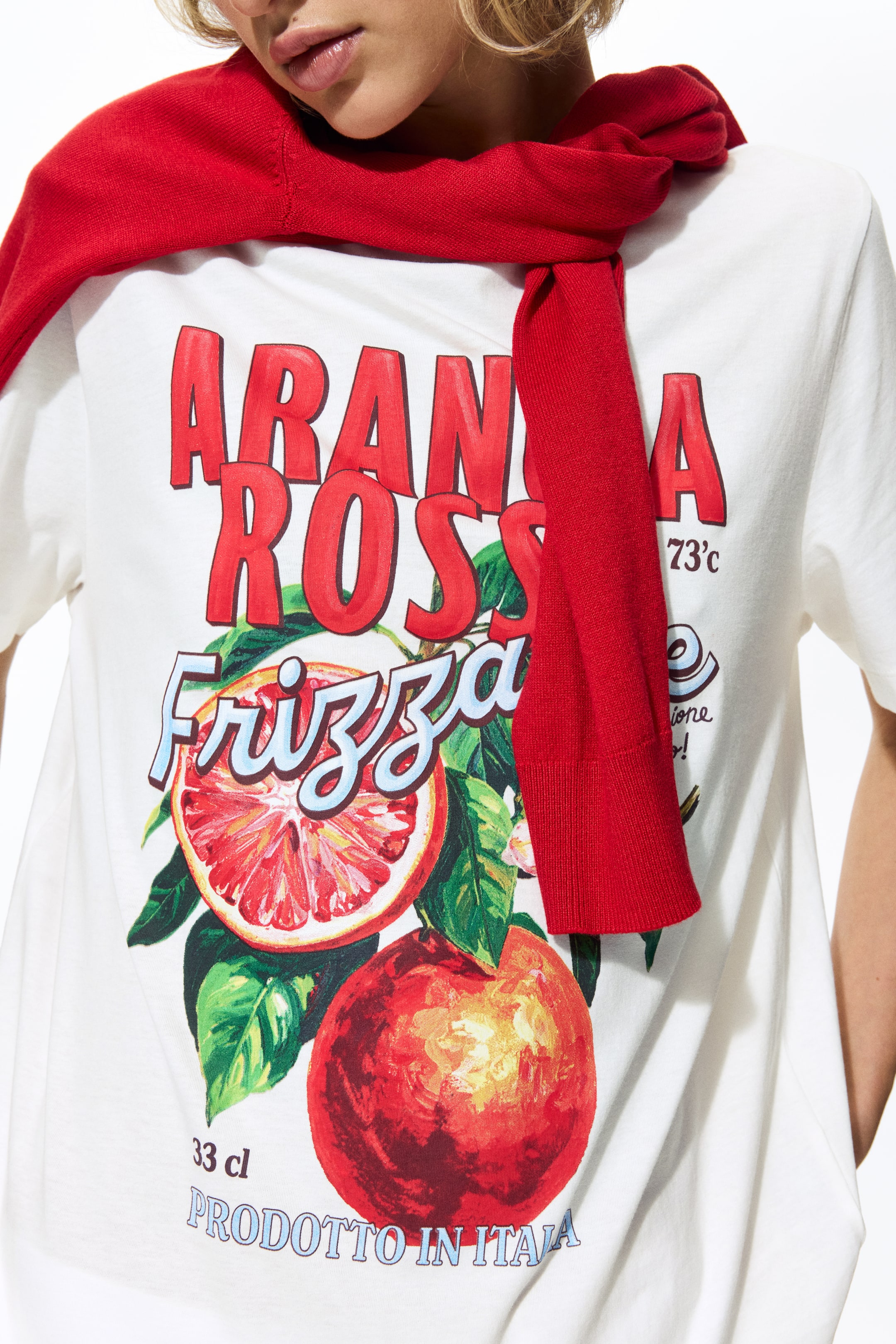 View larger image: A white short-sleeve t-shirt features a graphic print of "ARANCIA ROSSA Frizza" in red and blue text, accompanied by illustrations of blood oranges and leaves, with additional text "33 cl PRODOTTO IN ITALIA." A red sweater is draped over the shoulders, partially covering the upper part of the t-shirt.