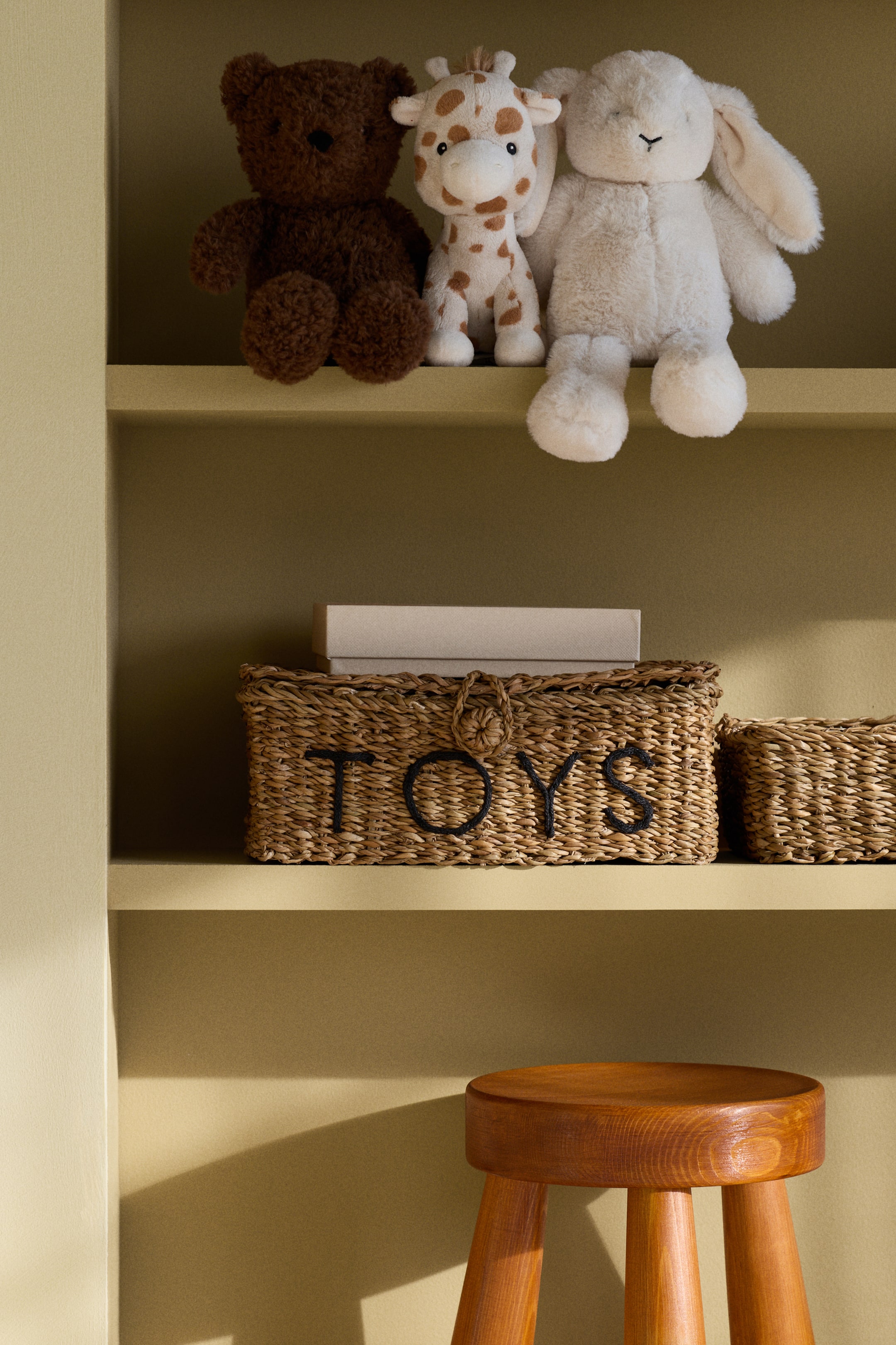 View larger image: Three plush toys are sitting facing forward: a dark brown teddy bear, a spotted light brown and white giraffe, and a cream bunny. A rectangular woven basket with 'TOYS' in dark letters and a loop closure is resting next to a smaller matching woven basket. A round wooden stool with three splayed legs is displayed.