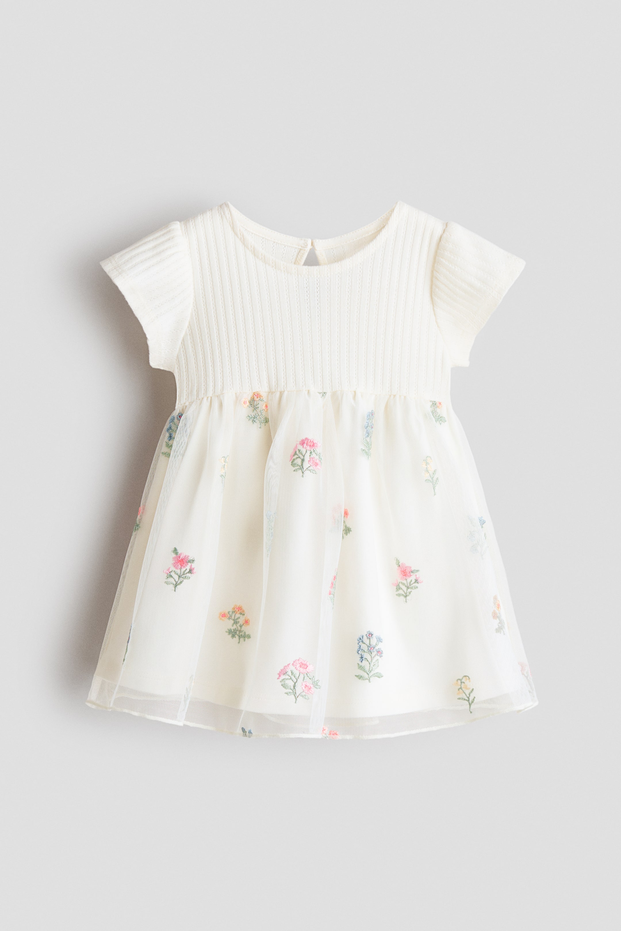 Ver imagen más grande: Un vestido de bebé presenta su PARTE DELANTERA, con un cuerpo de punto acanalado color crema con escote redondo y mangas cortas. Su falda acampanada muestra un forro color crema cubierto por tul transparente, bordado con motivos florales coloridos y dispersos.